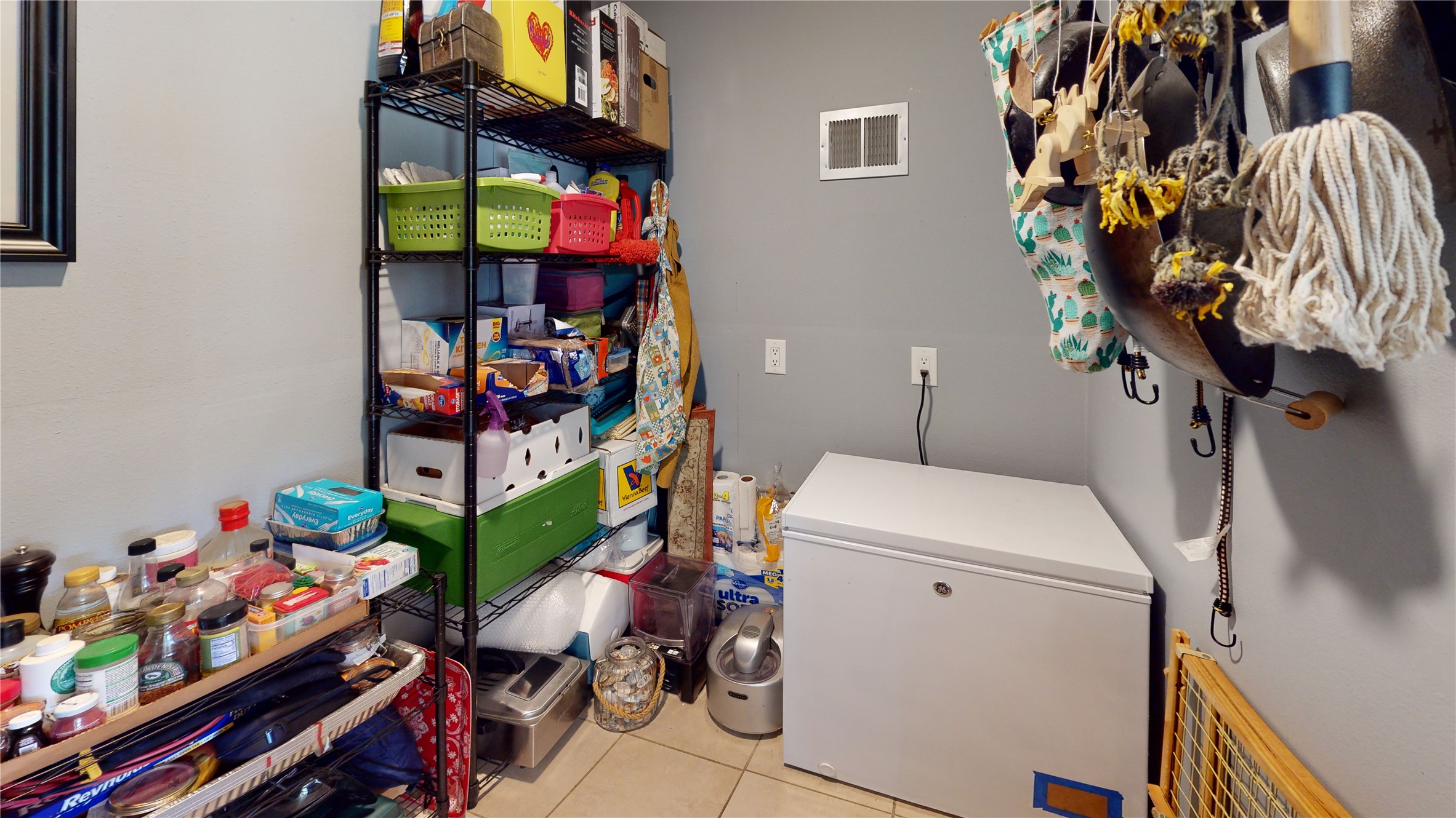 Utility storage room with a chest freezer, crowded shelving, and hanging household items.