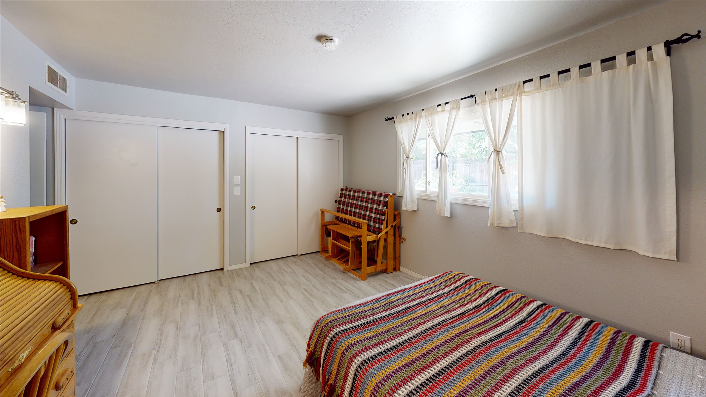 Bedroom with a colorful striped blanket, pale tile floor, sliding closets, and a curtained window.