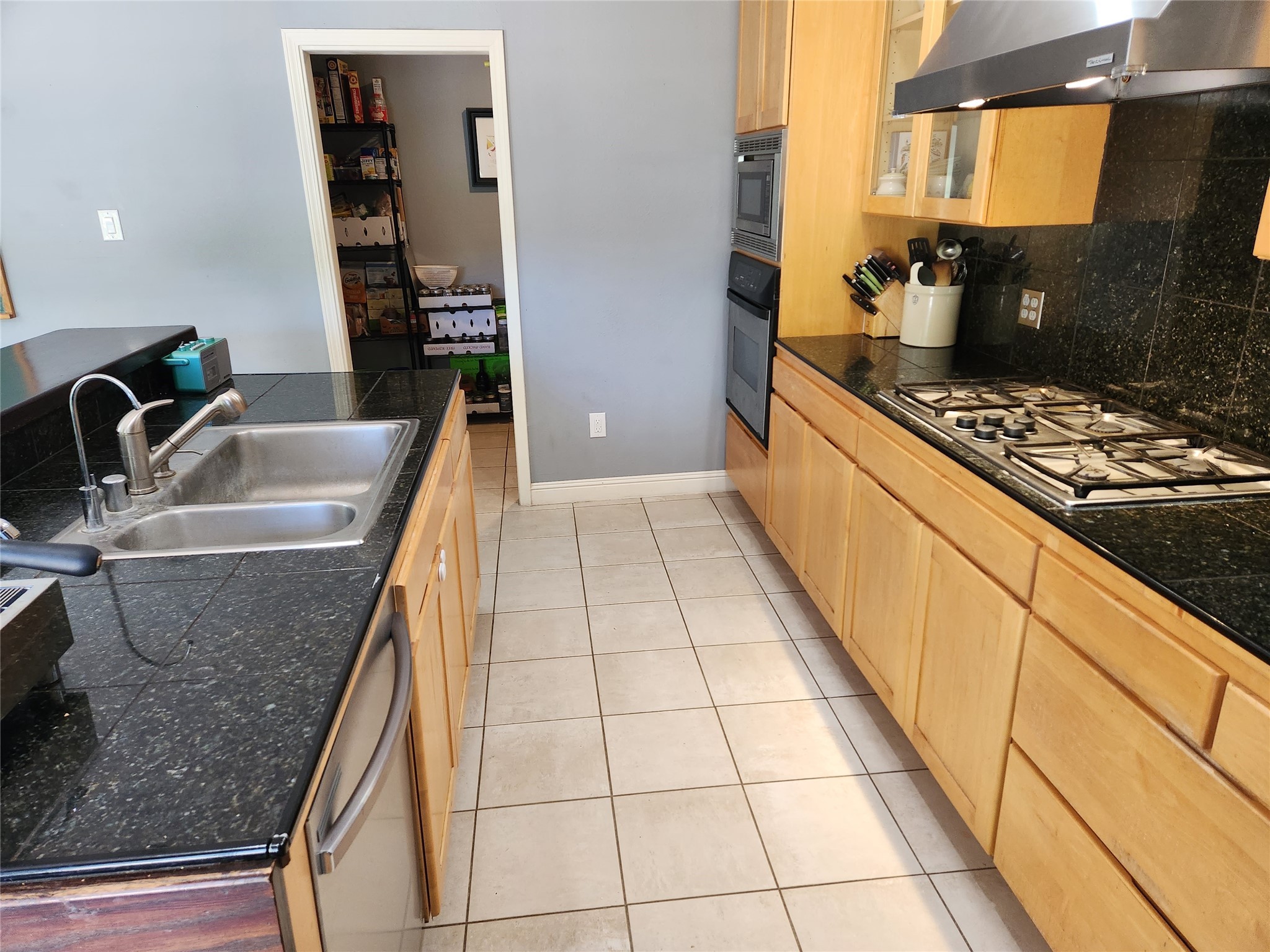 Kitchen with light wood cabinets, black counters, a gas cooktop, double sink, and stocked pantry doorway.