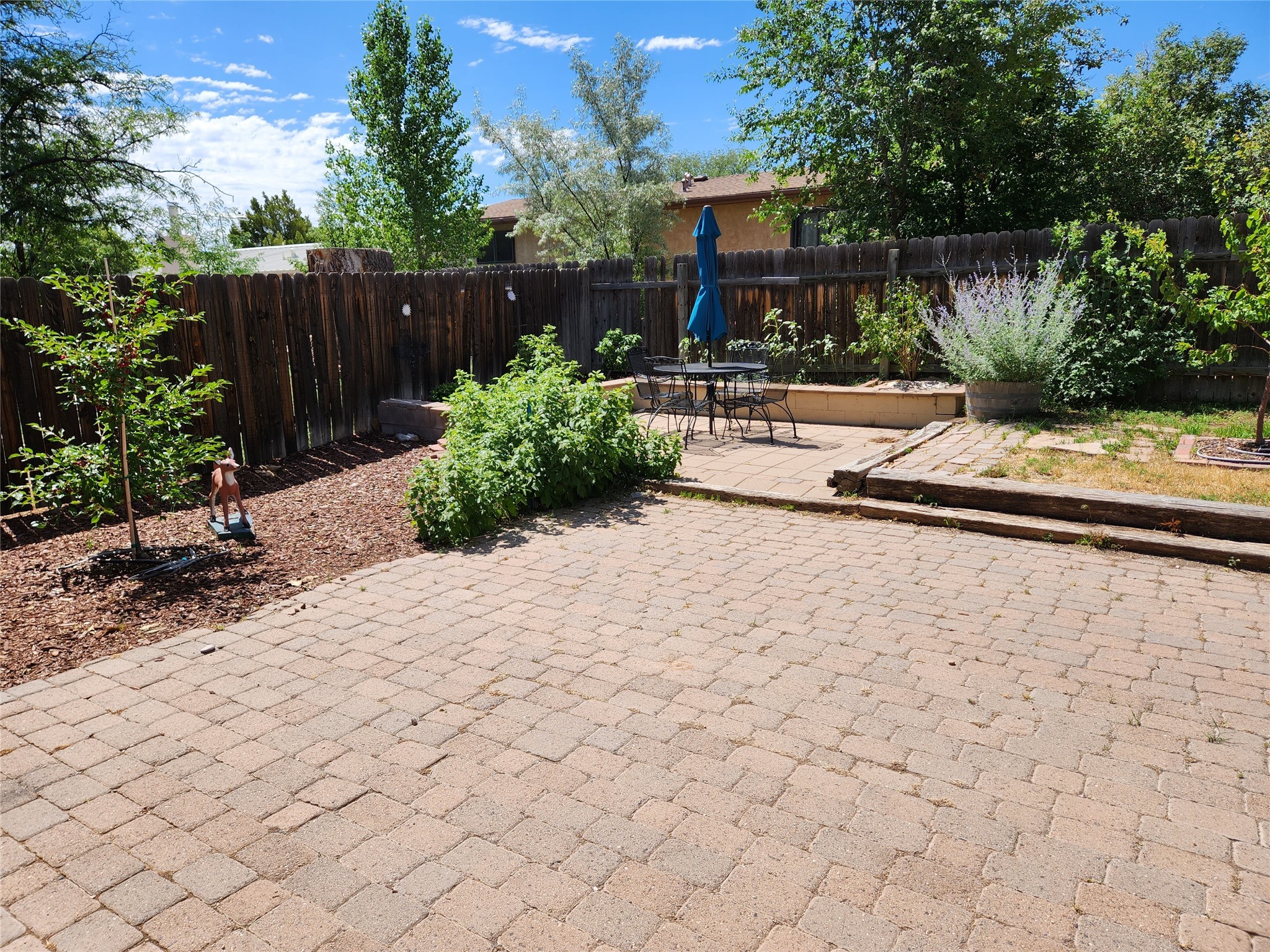 Backyard patio with pavers, outdoor dining set, blue umbrella, raised beds, and a wooden fence.