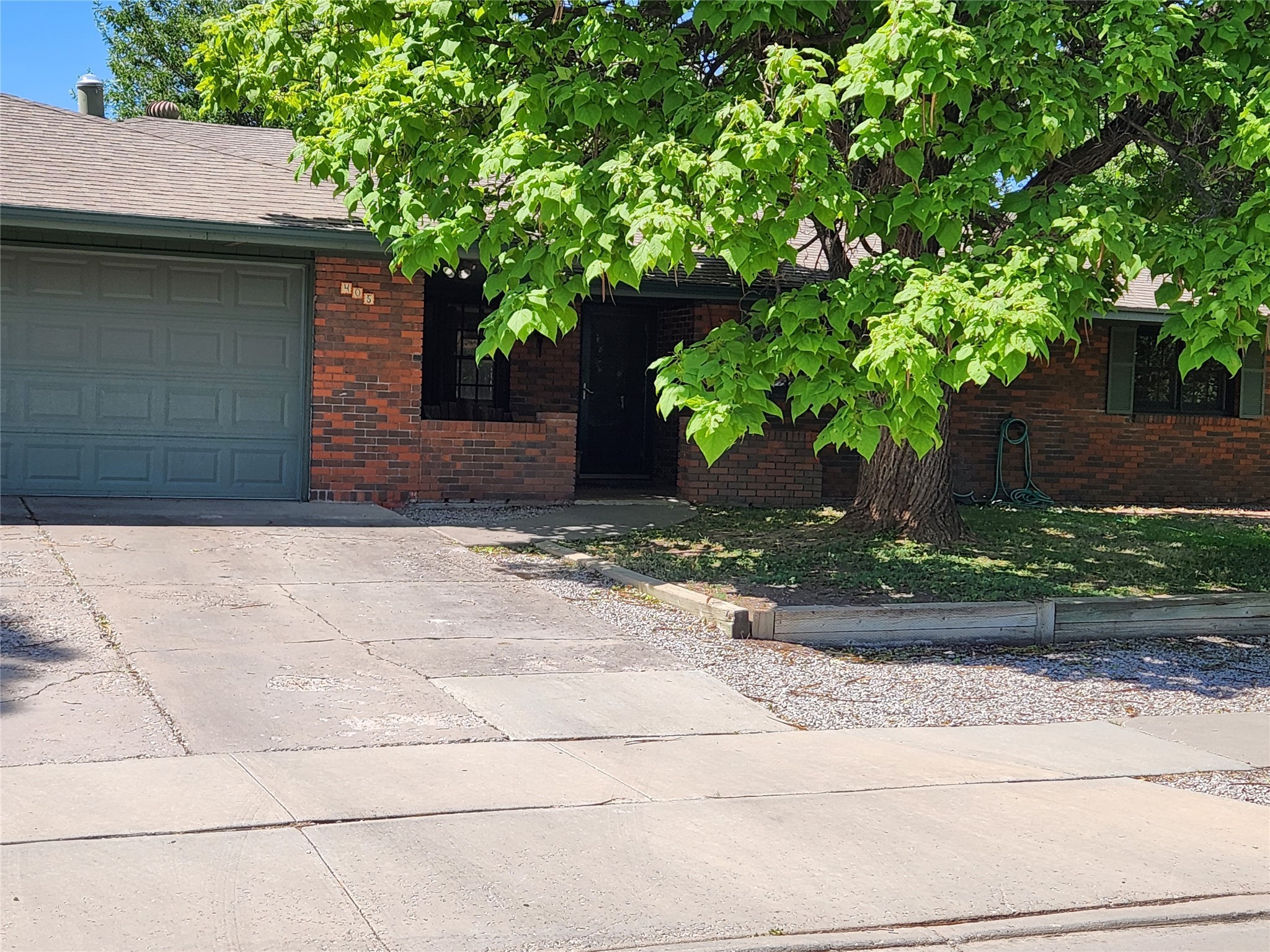 Red brick house with a gray garage door and large leafy tree shading the driveway.