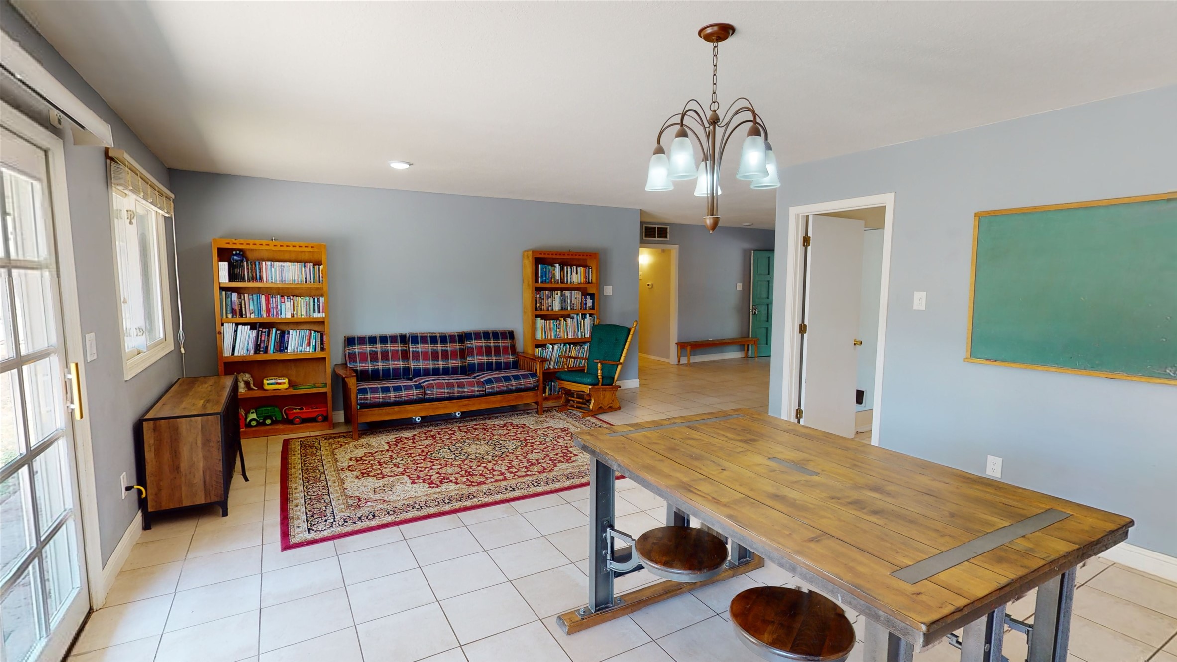 Open dining and living area with a long wooden table, plaid sofa, bookshelves, and green chalkboard.