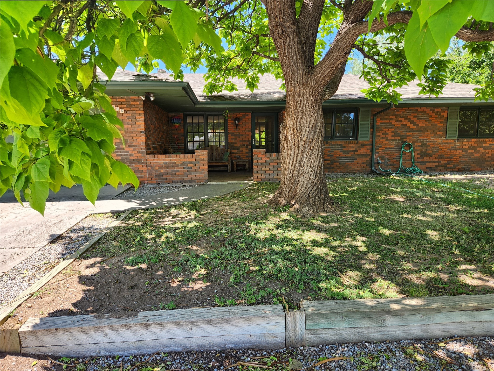 Single-story brick home shaded by a large tree, with a recessed entry and timber-bordered yard.