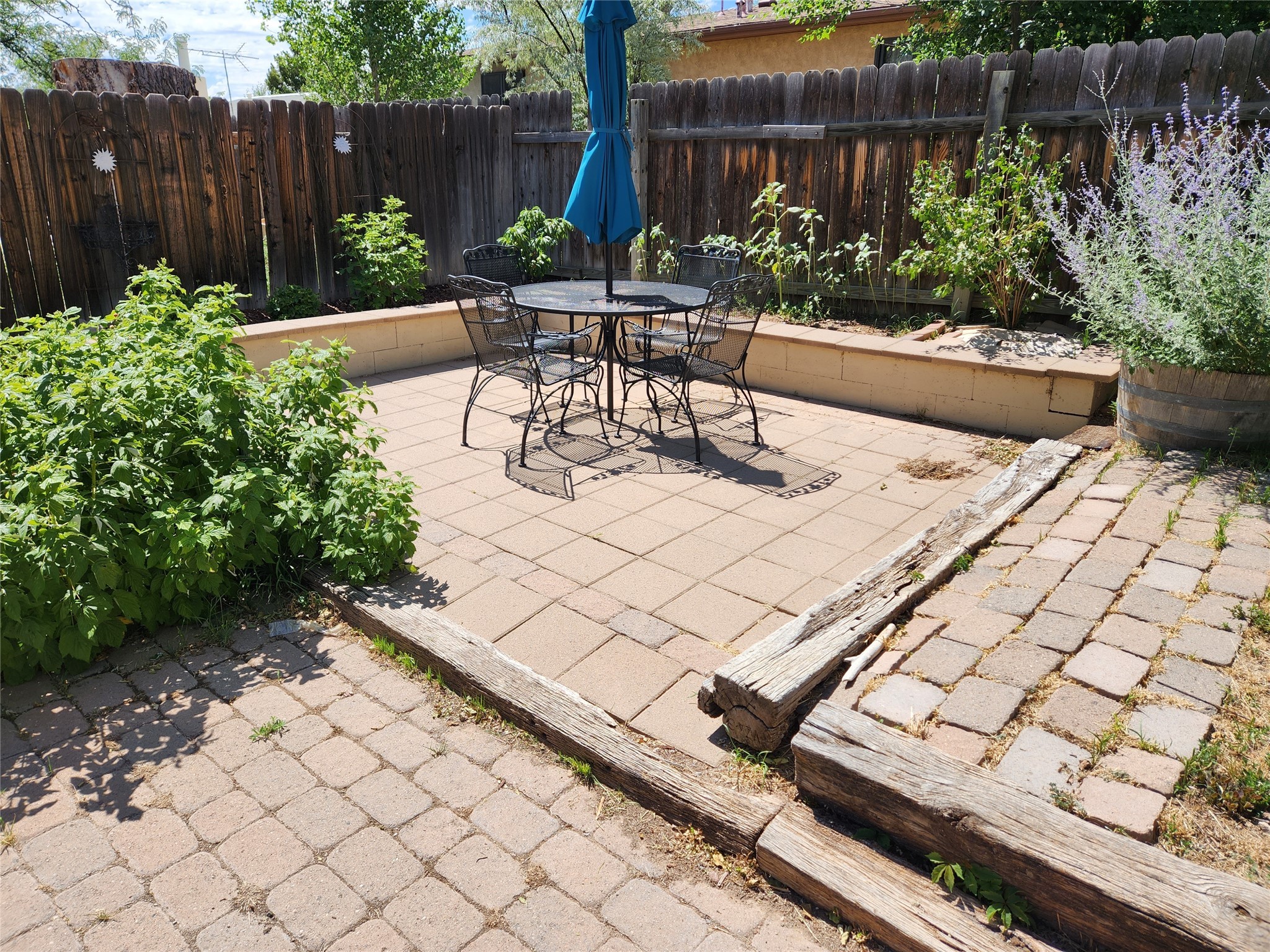 Fenced paver patio with a metal dining set, blue umbrella, and raised garden beds.