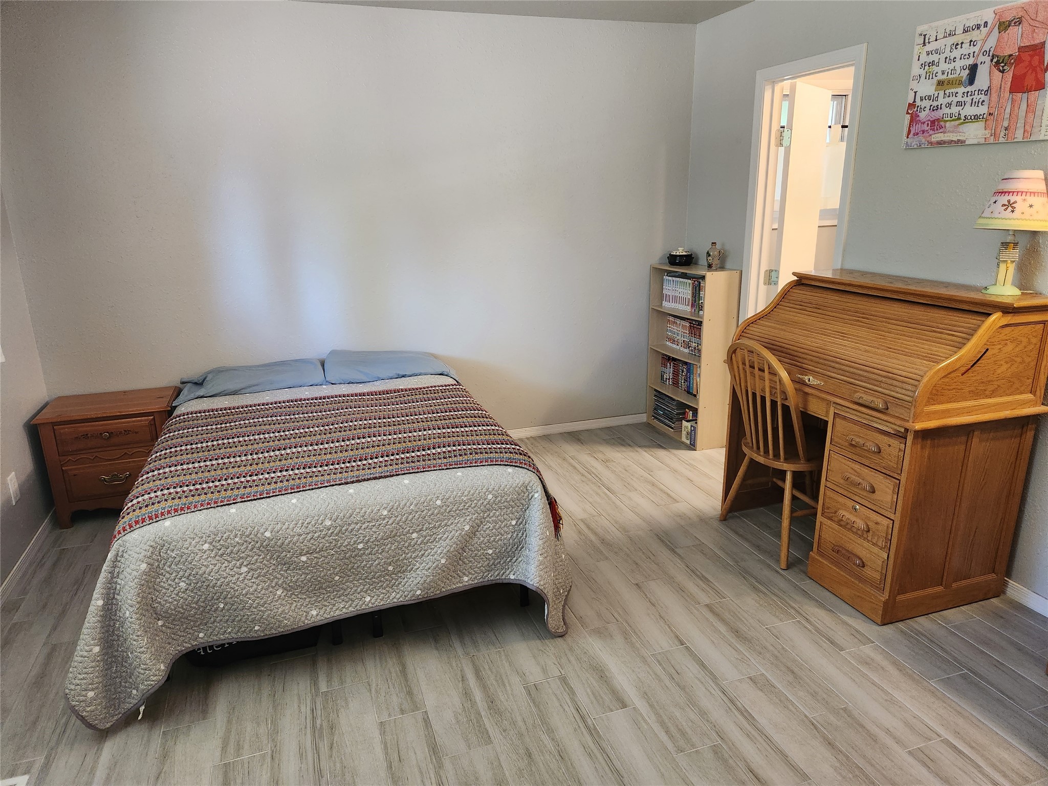 Bedroom with a patterned bedspread, wooden roll-top desk, and open doorway to a bright adjoining room.