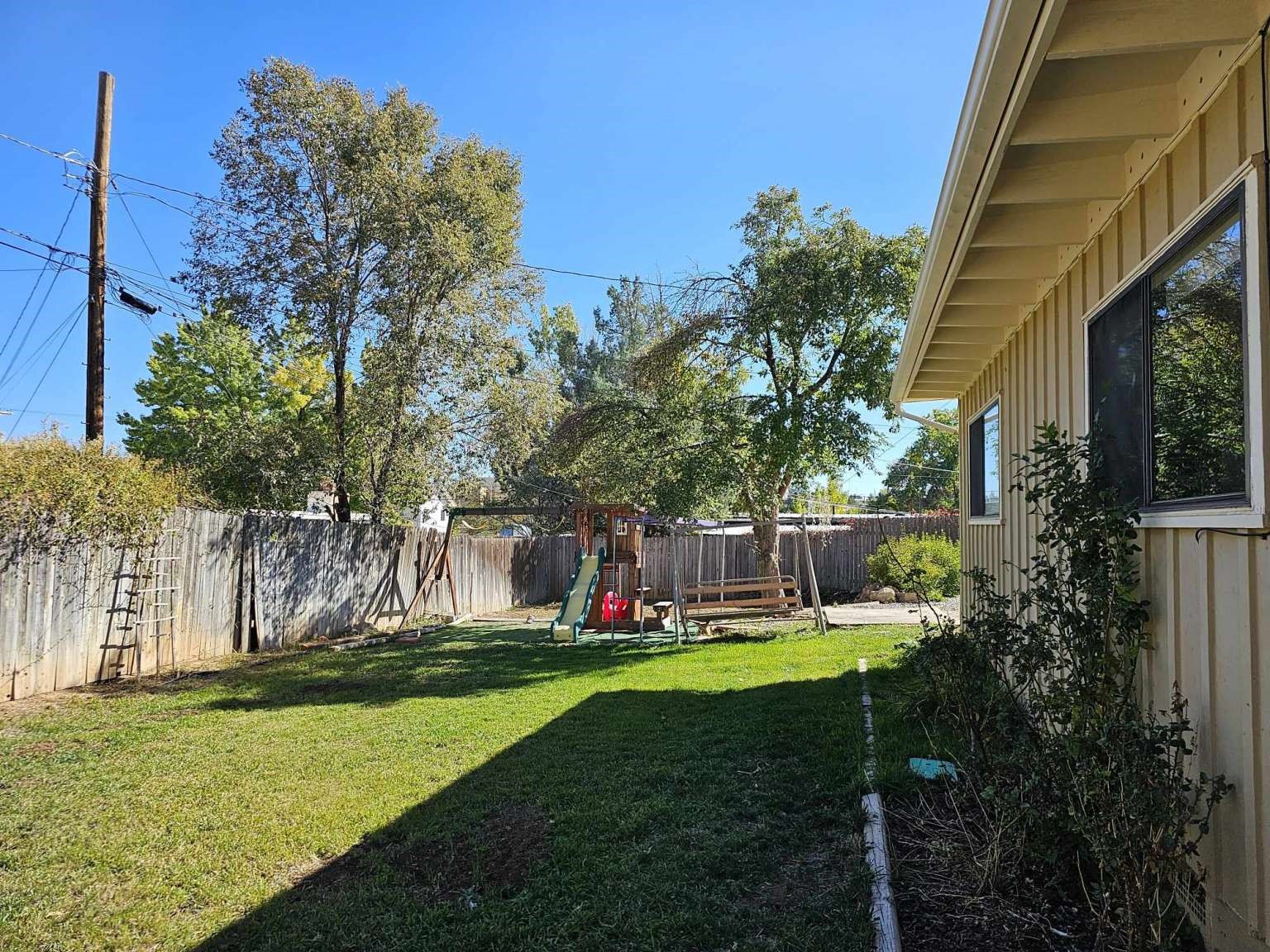 Fenced backyard with a lawn, mature trees, and a children's swing set beside the house.