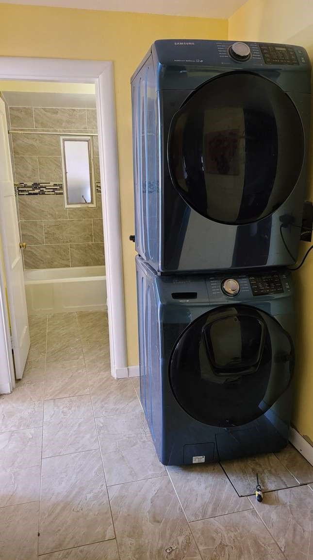 Laundry room with stacked dark front-loading washer and dryer beside a tiled bathroom doorway.