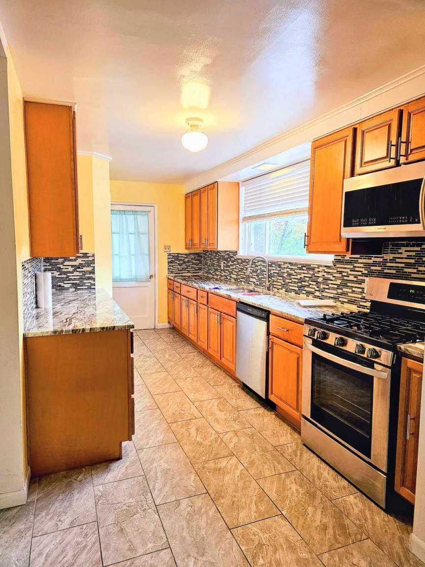 Galley kitchen with warm wood cabinets, mosaic tile backsplash, stainless steel appliances, and a window above the sink.