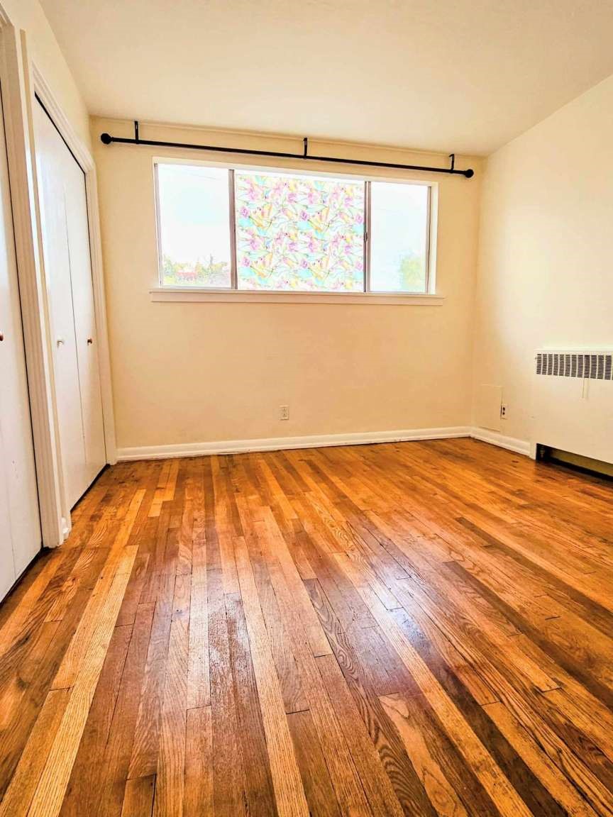 Empty bedroom with polished hardwood floors, white closet doors, and a wide window.