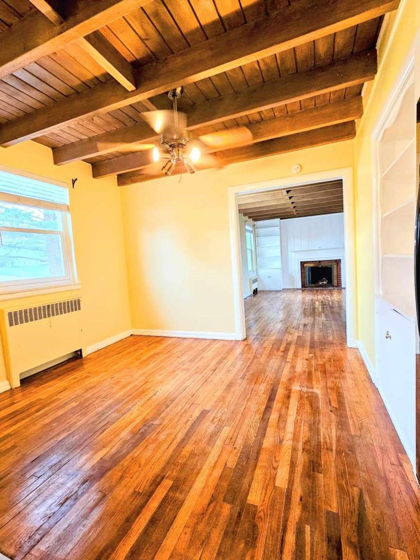 Empty room with polished hardwood floors and exposed wood beams, opening to a room with a fireplace.