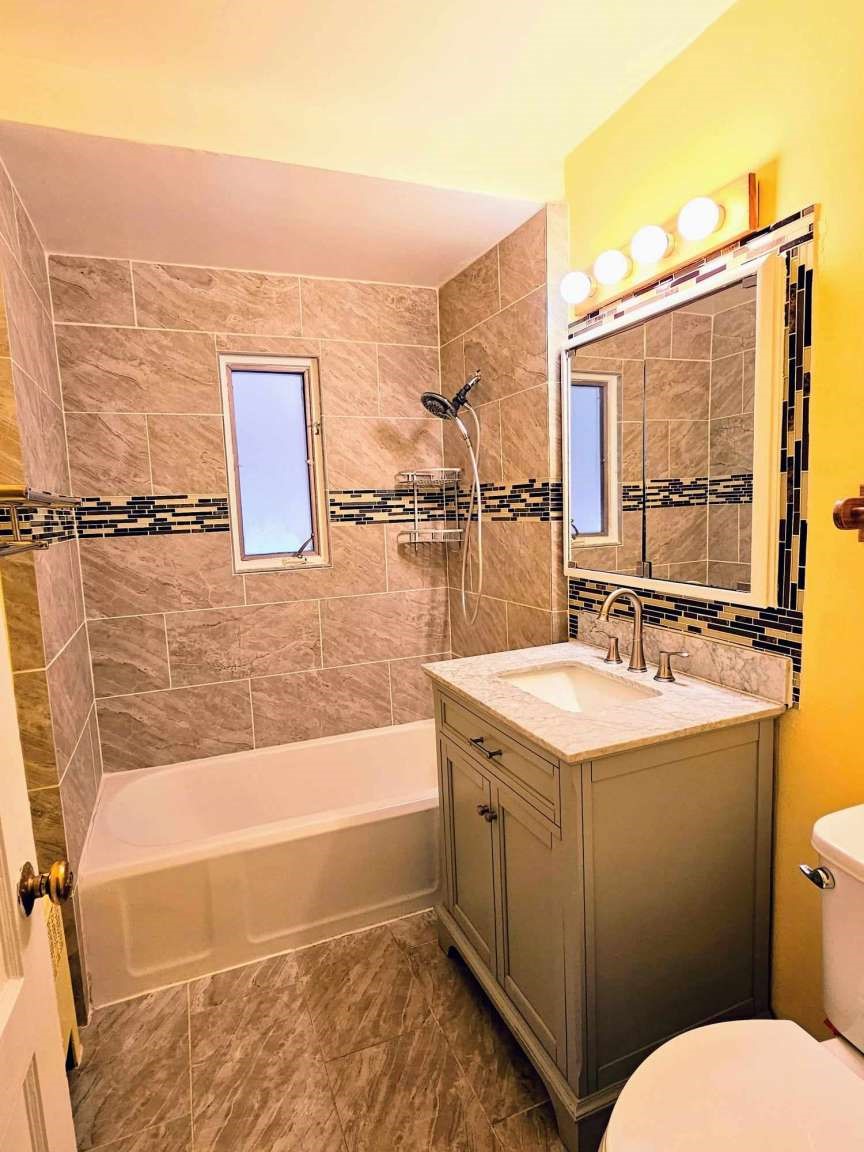 Bathroom with a tiled tub and shower, narrow window, and gray vanity beneath a large mirror.
