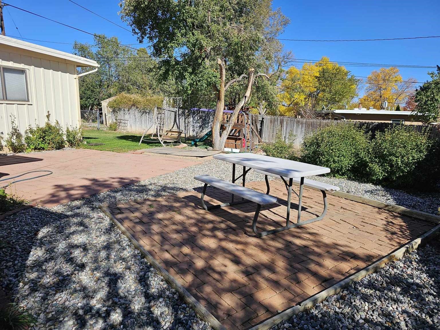 Backyard patio with brick pavers, picnic table, gravel landscaping, and a fenced play area.