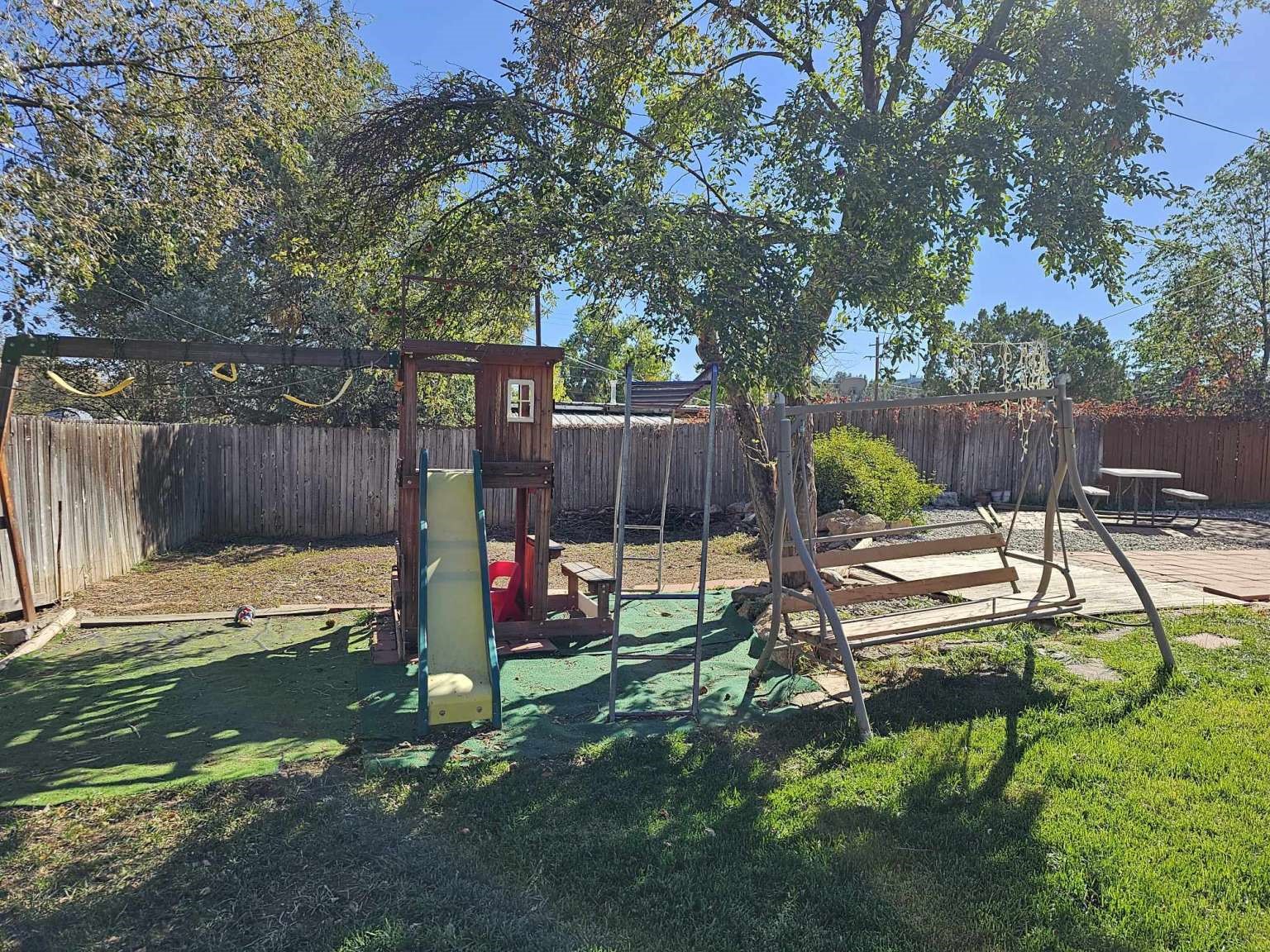 Fenced backyard with a wooden play structure, slide, swing set, and picnic table beneath a large tree.