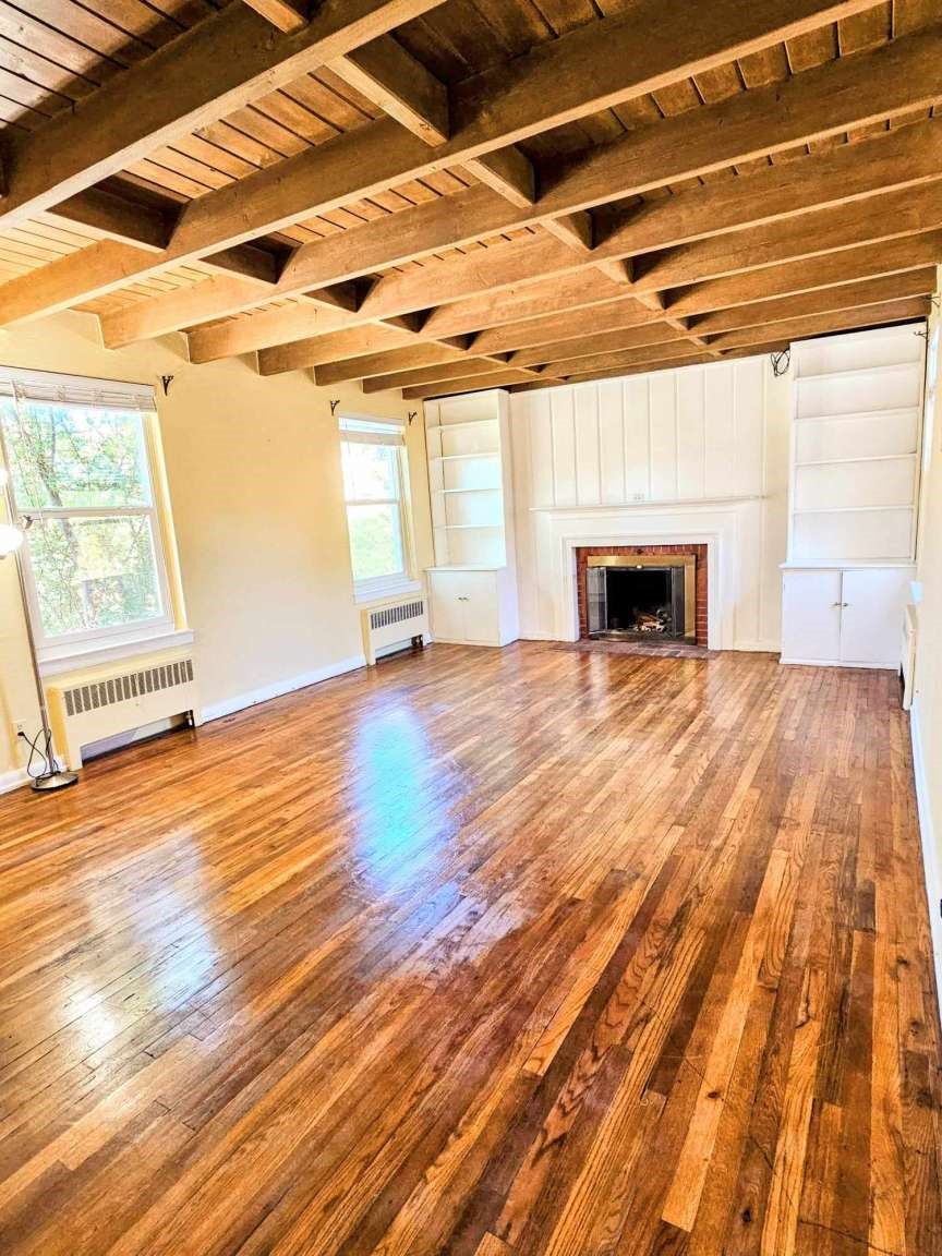 Empty living room with exposed wood beams, polished hardwood floors, built-in shelves, and a fireplace.