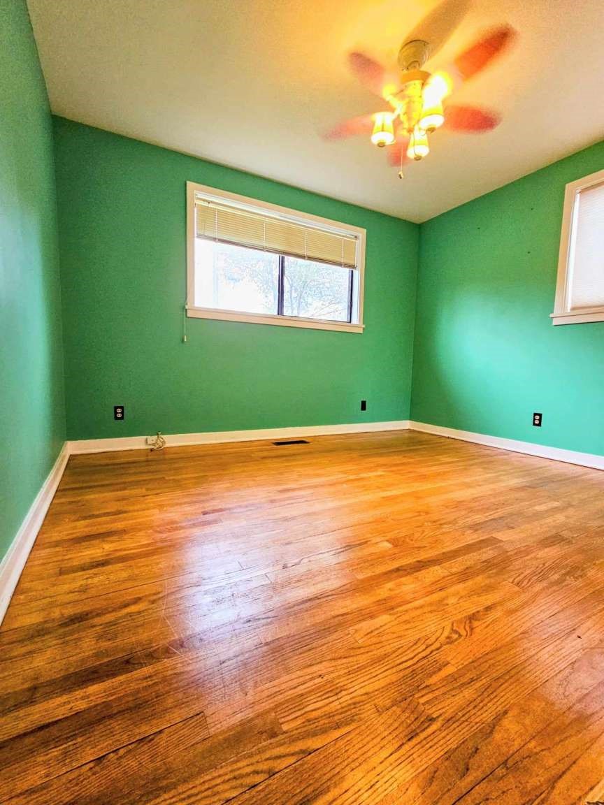 Empty bedroom with green walls, polished hardwood floors, and a ceiling fan with lights.