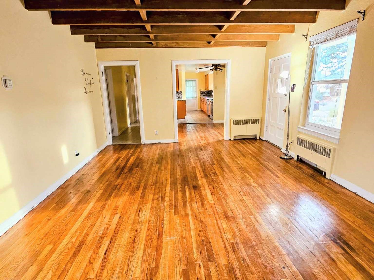 Empty living room with polished hardwood floors, exposed ceiling beams, and a view into the kitchen.