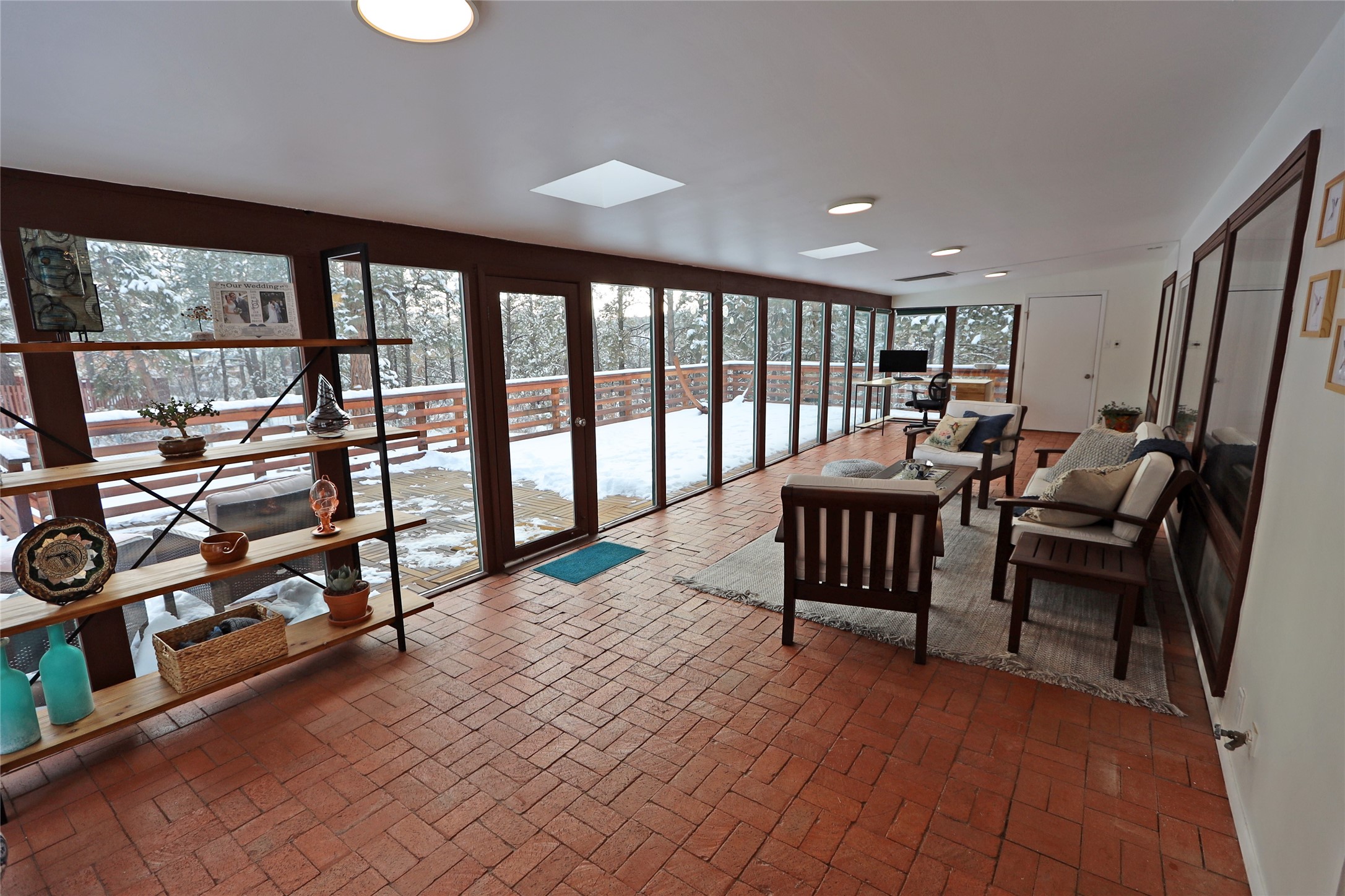 Furnished sunroom with brick flooring and glass doors overlooking a snow-covered deck and trees.