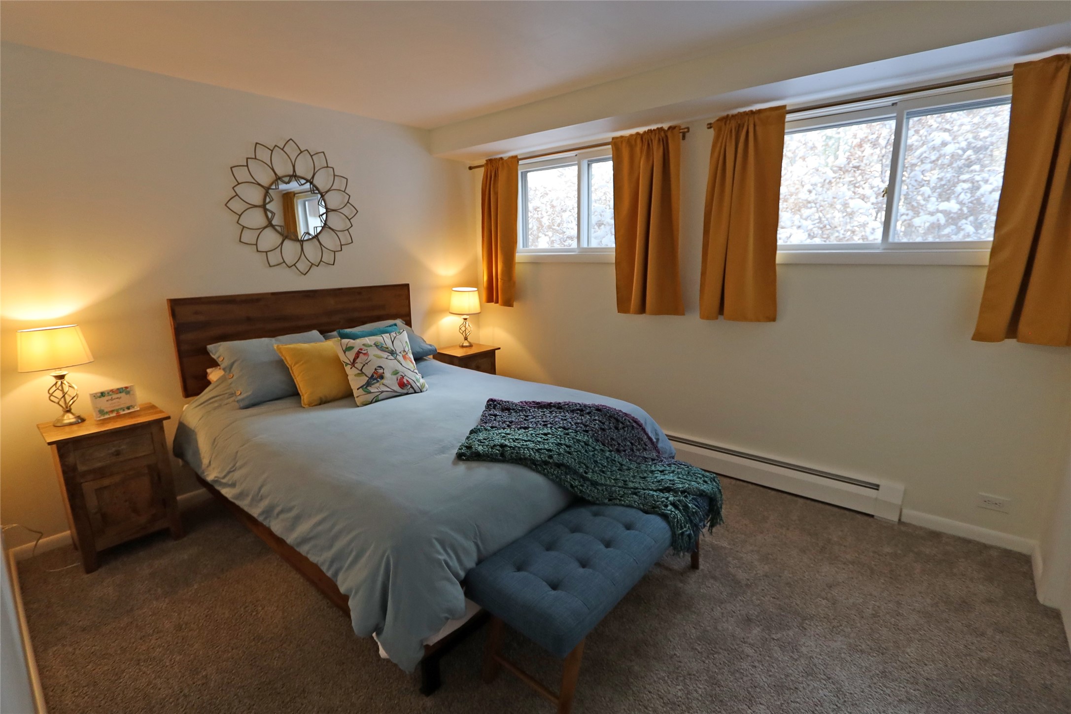 Furnished bedroom with a wooden bed, blue bench, rust-colored curtains, and snowy trees visible through the windows.
