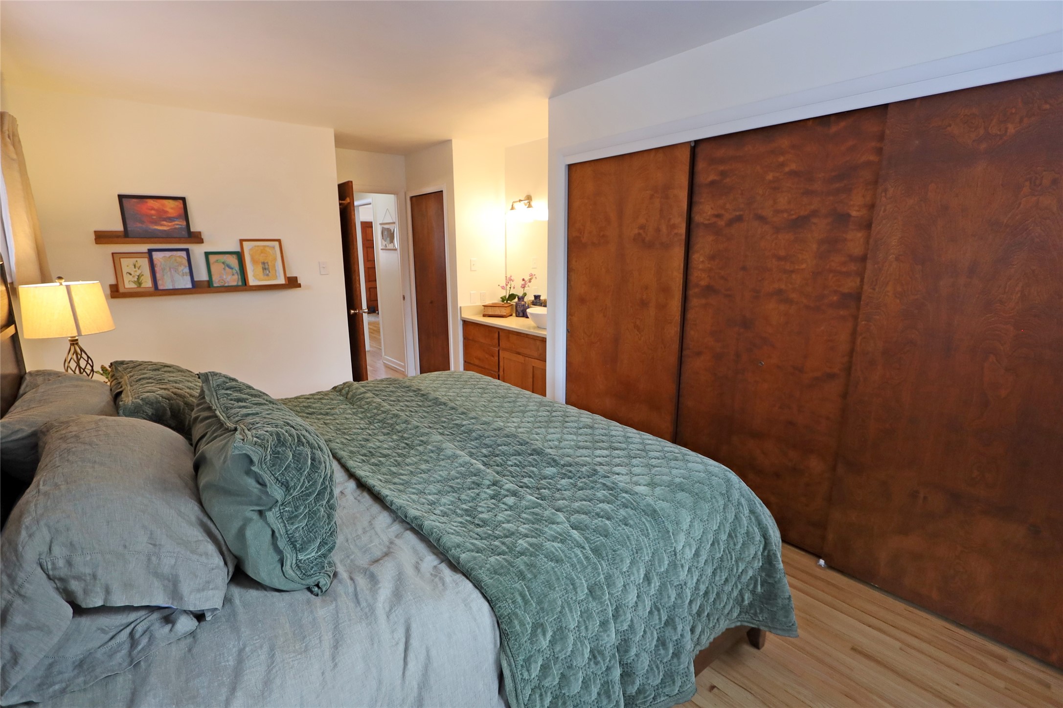 Bedroom with a sage-green quilt, wood sliding closet doors, and framed artwork on wall shelves.