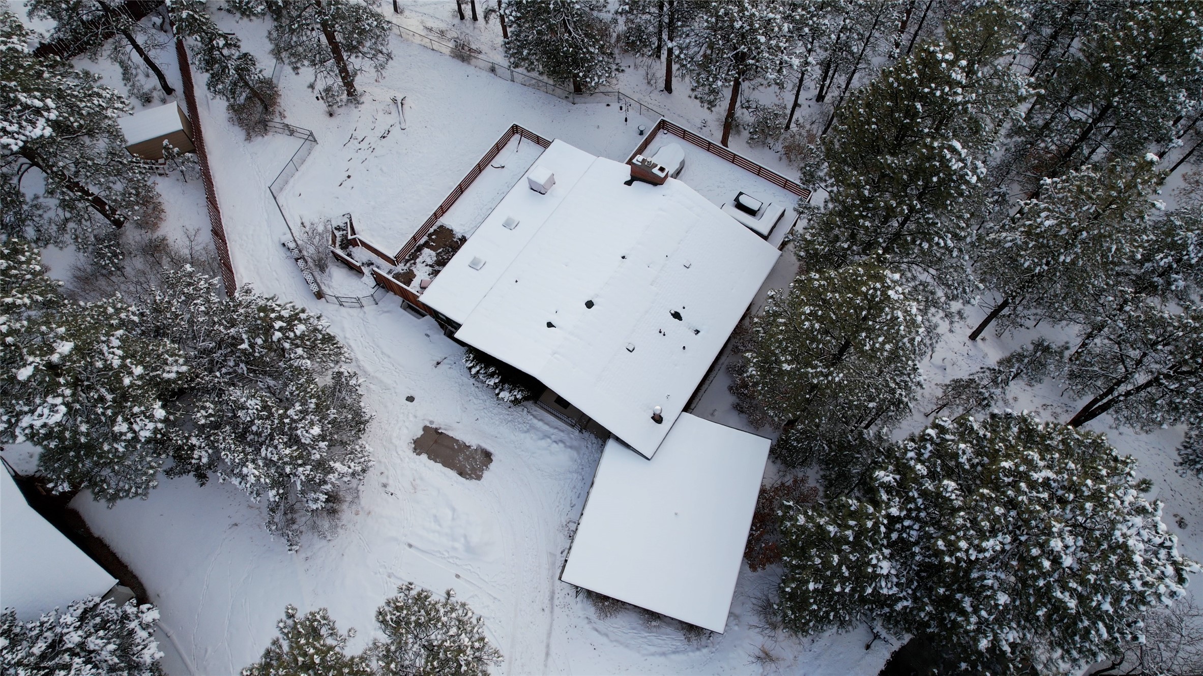 Snow-covered home and fenced deck among trees, viewed from above.