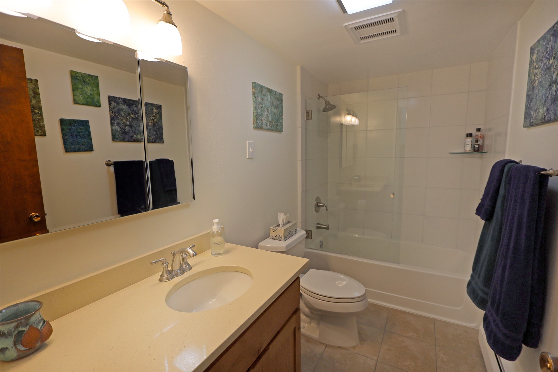 Bathroom with a cream vanity, mirrored cabinets, and a white-tiled tub-shower with glass doors.