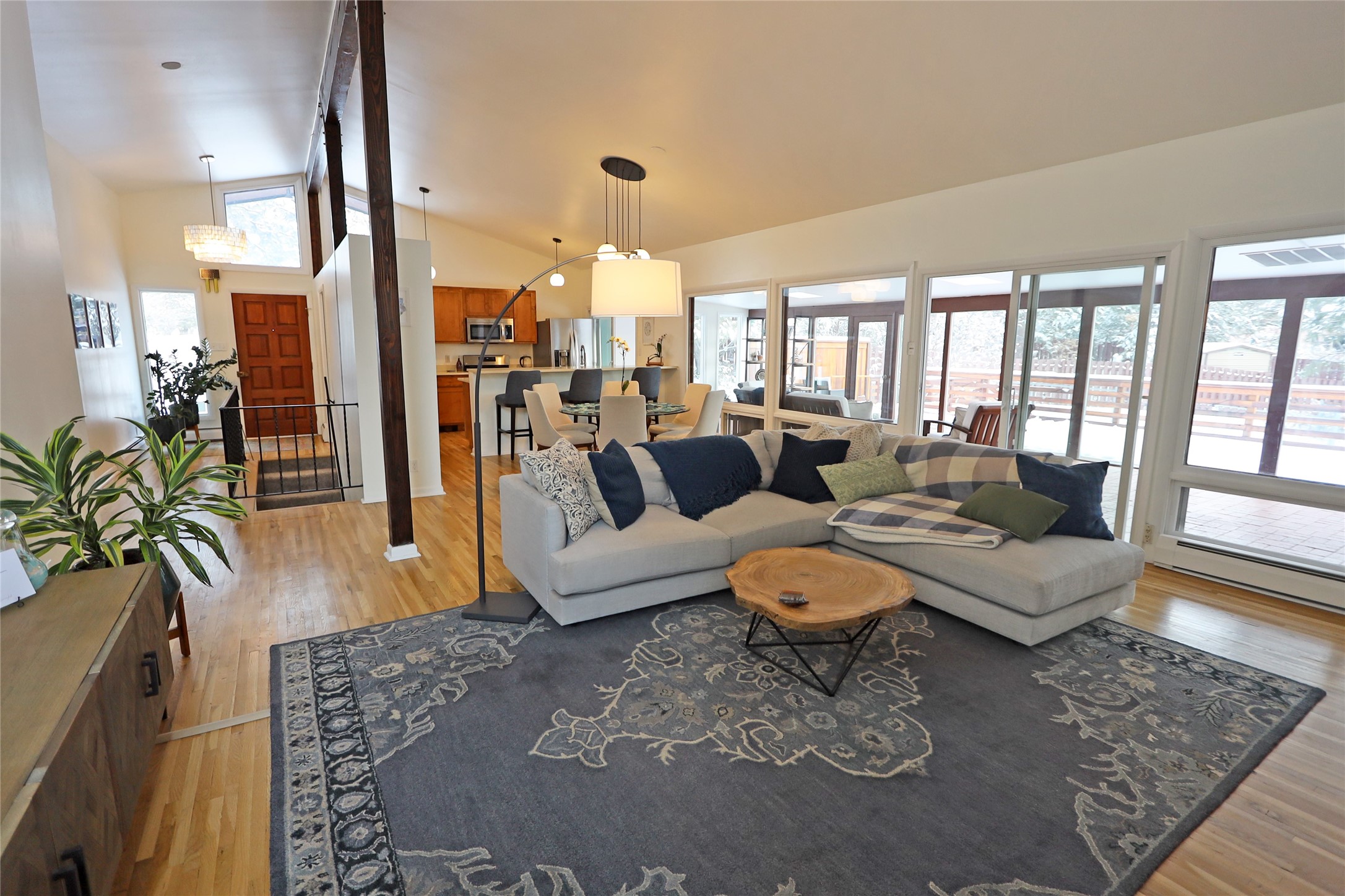 Open-plan living room with a gray sectional, patterned rug, and large windows overlooking a snowy deck.