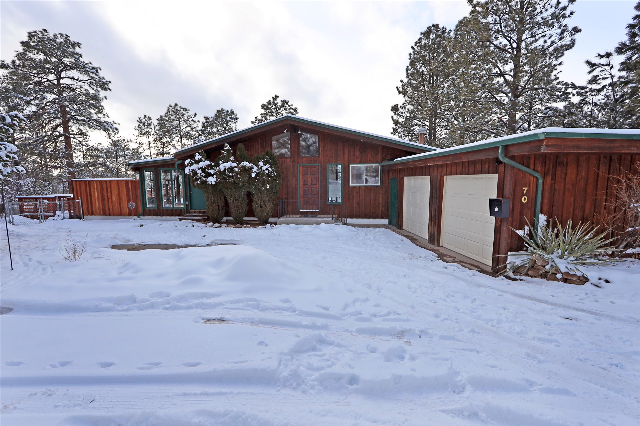 Wood-sided home with green trim and attached garage, surrounded by snow-covered ground and pine trees.