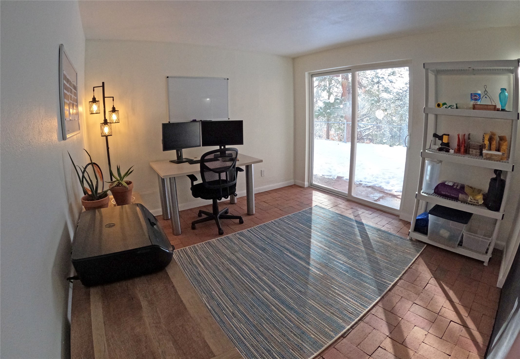 Home office with a dual-monitor desk beside a sliding glass door overlooking a snowy yard.