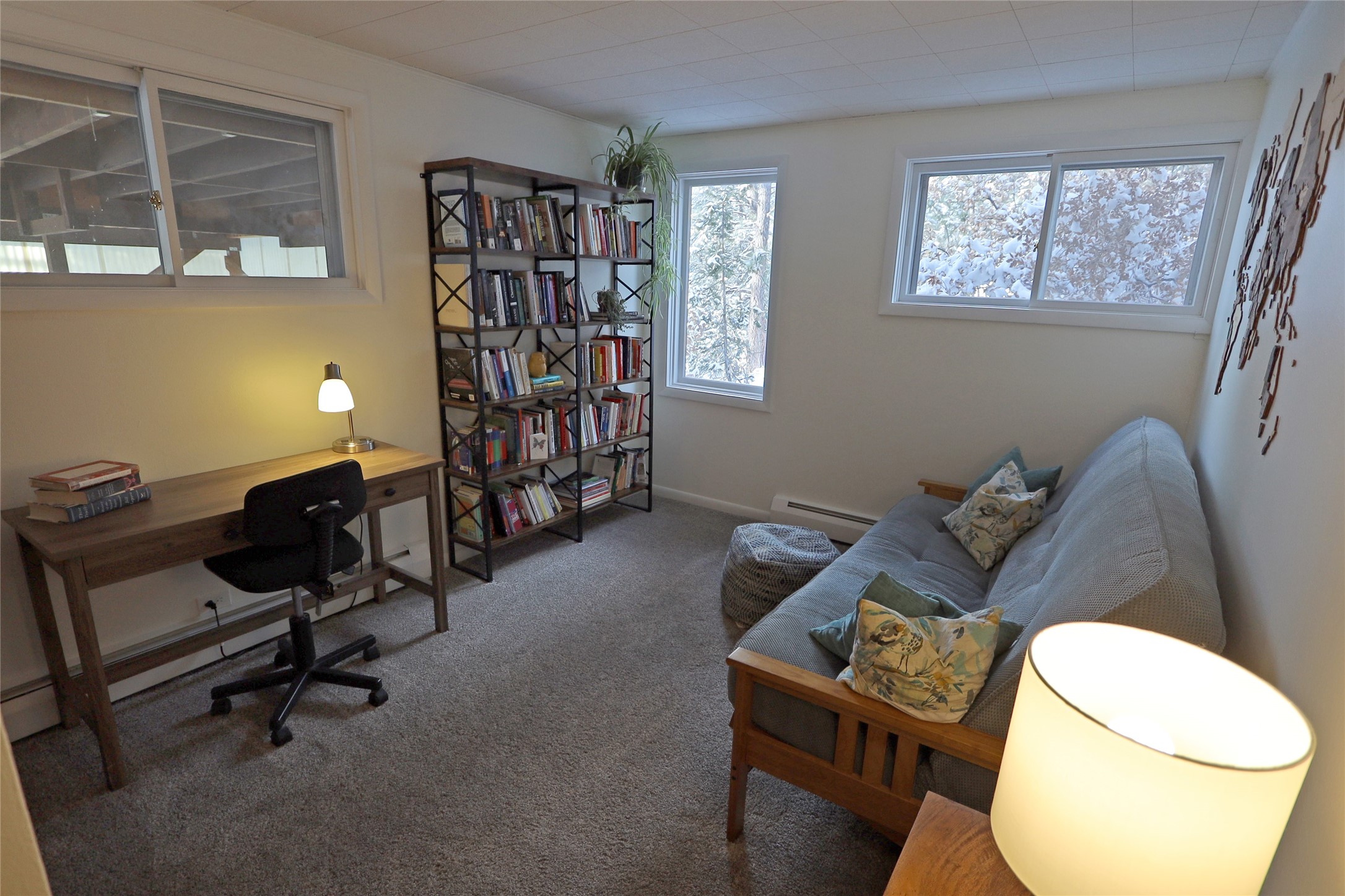 Home office with a wooden desk and bookcase beside a gray sofa and snowy windows.