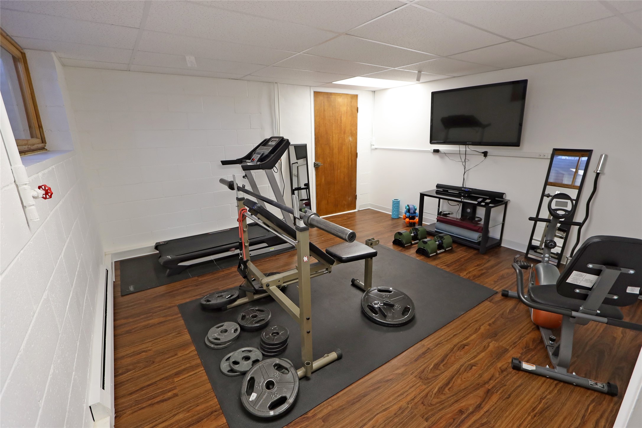 Basement gym with a weight bench, treadmill, elliptical machine, and wall-mounted television.