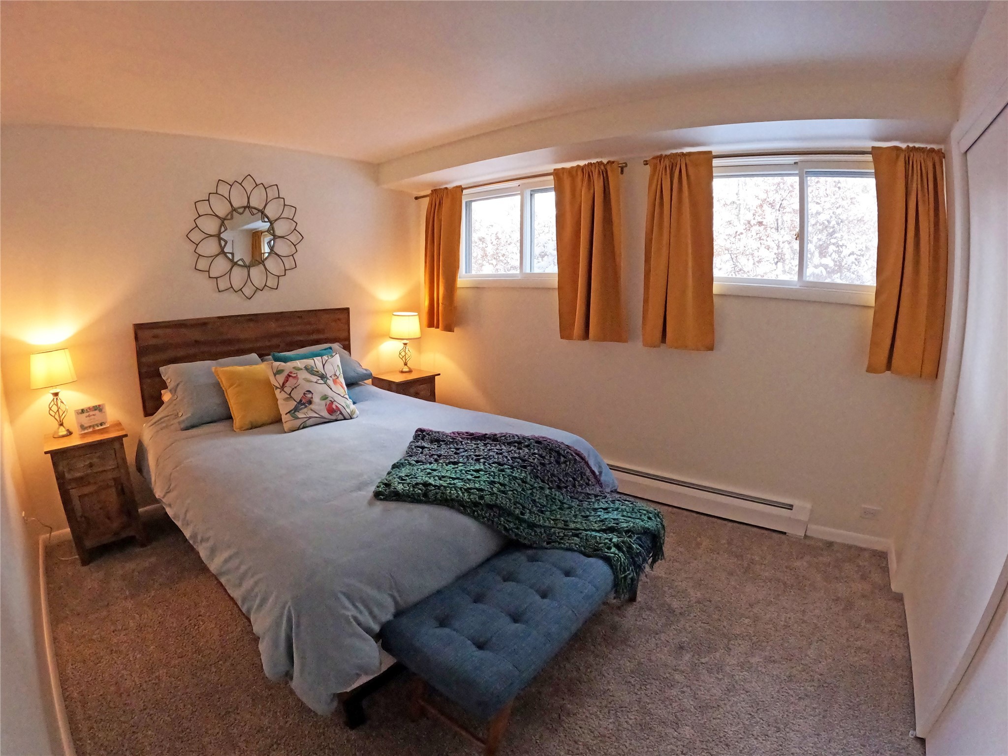 Staged bedroom with a gray bed, colorful pillows, knitted throw, and orange curtains framing high windows.