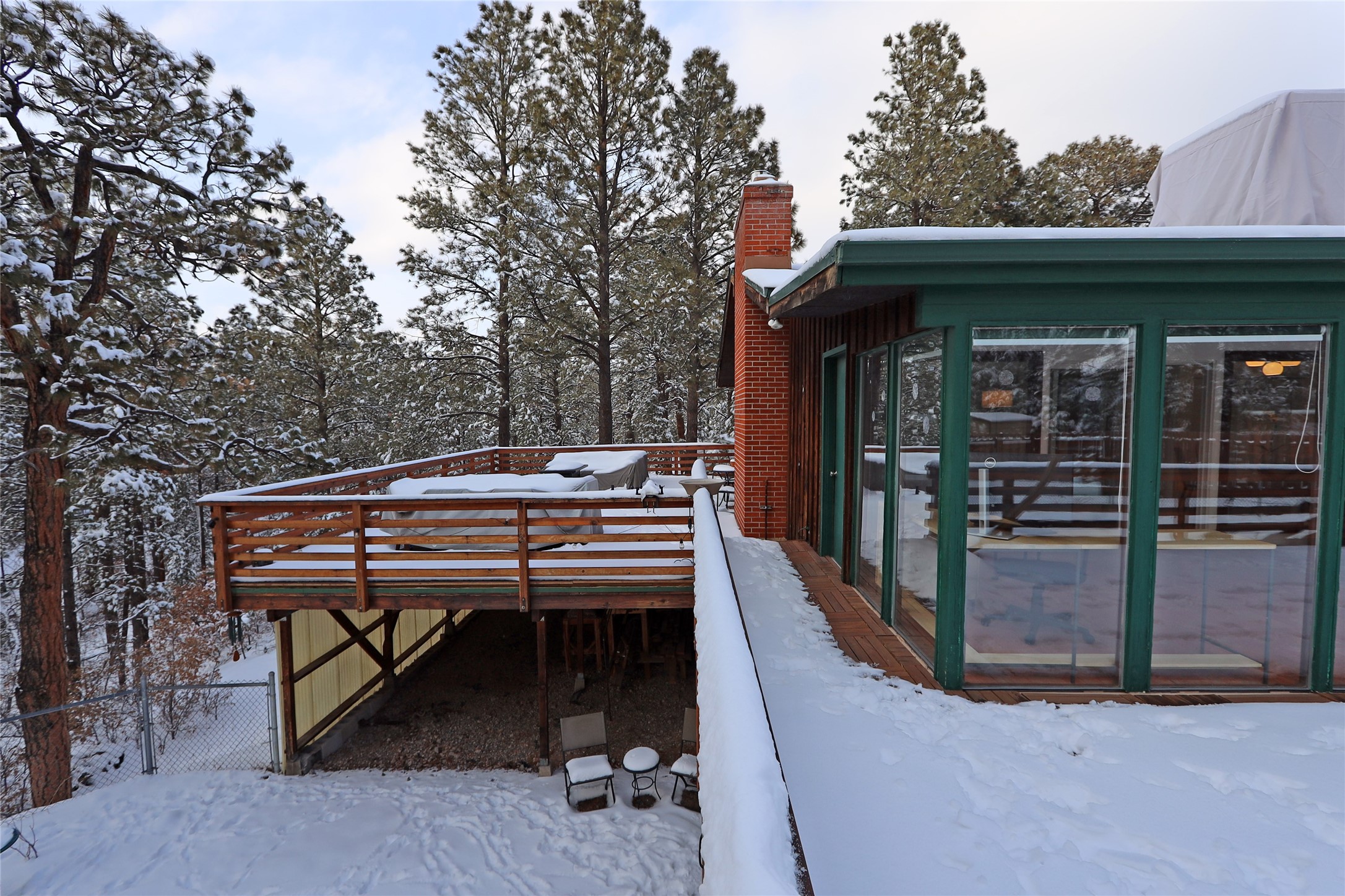 Snow-covered deck beside a glass sunroom, overlooking a snowy pine forest.