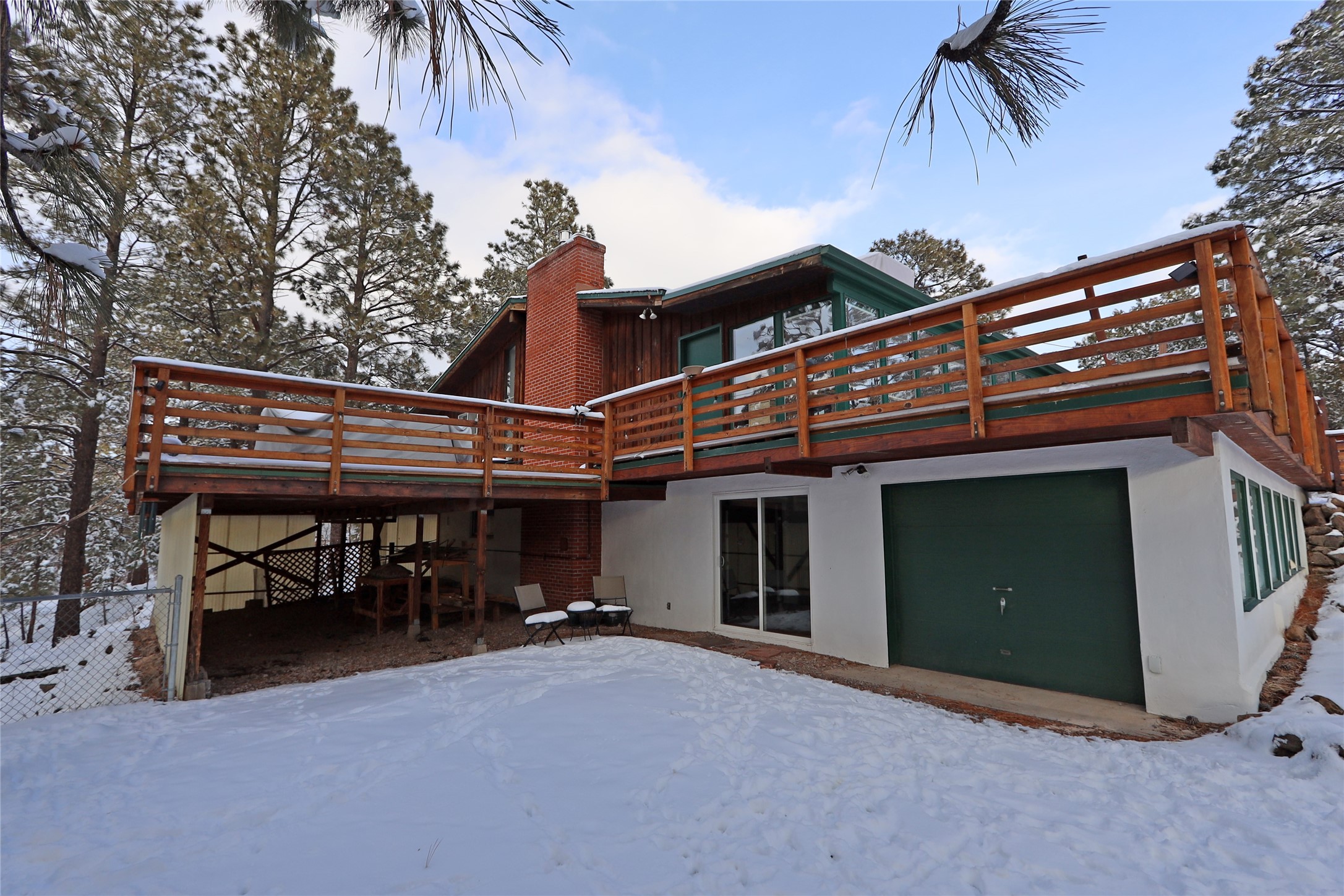 Snow-covered home with a large raised wooden deck, brick chimney, and green garage door.