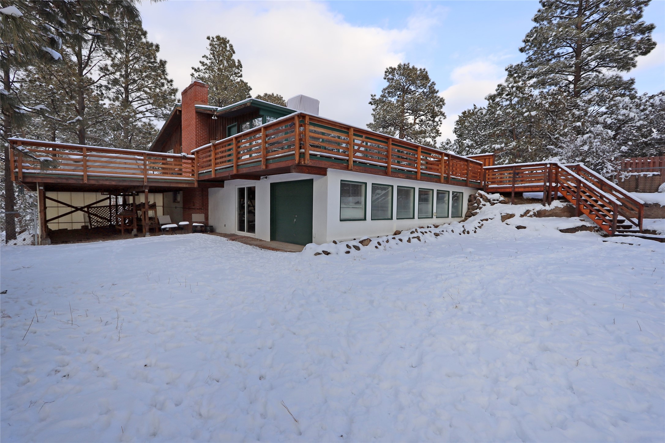 Snow-covered home with a broad elevated wooden deck and stairs among pine trees.