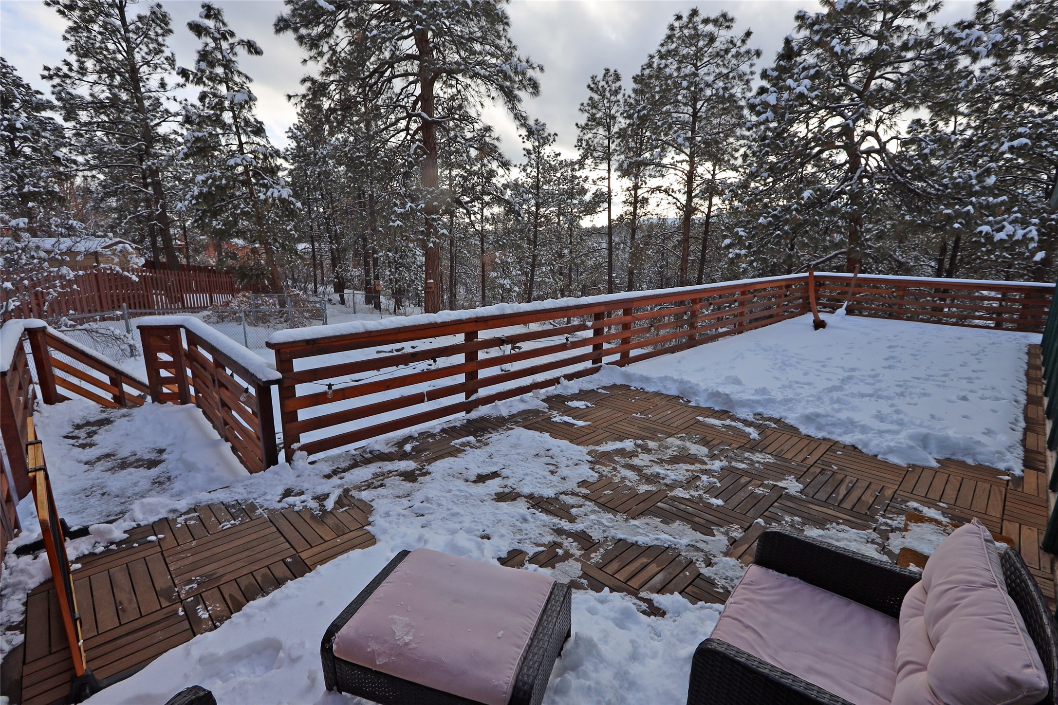 Snow-covered wooden deck with wicker seating and pine trees beyond the railing.