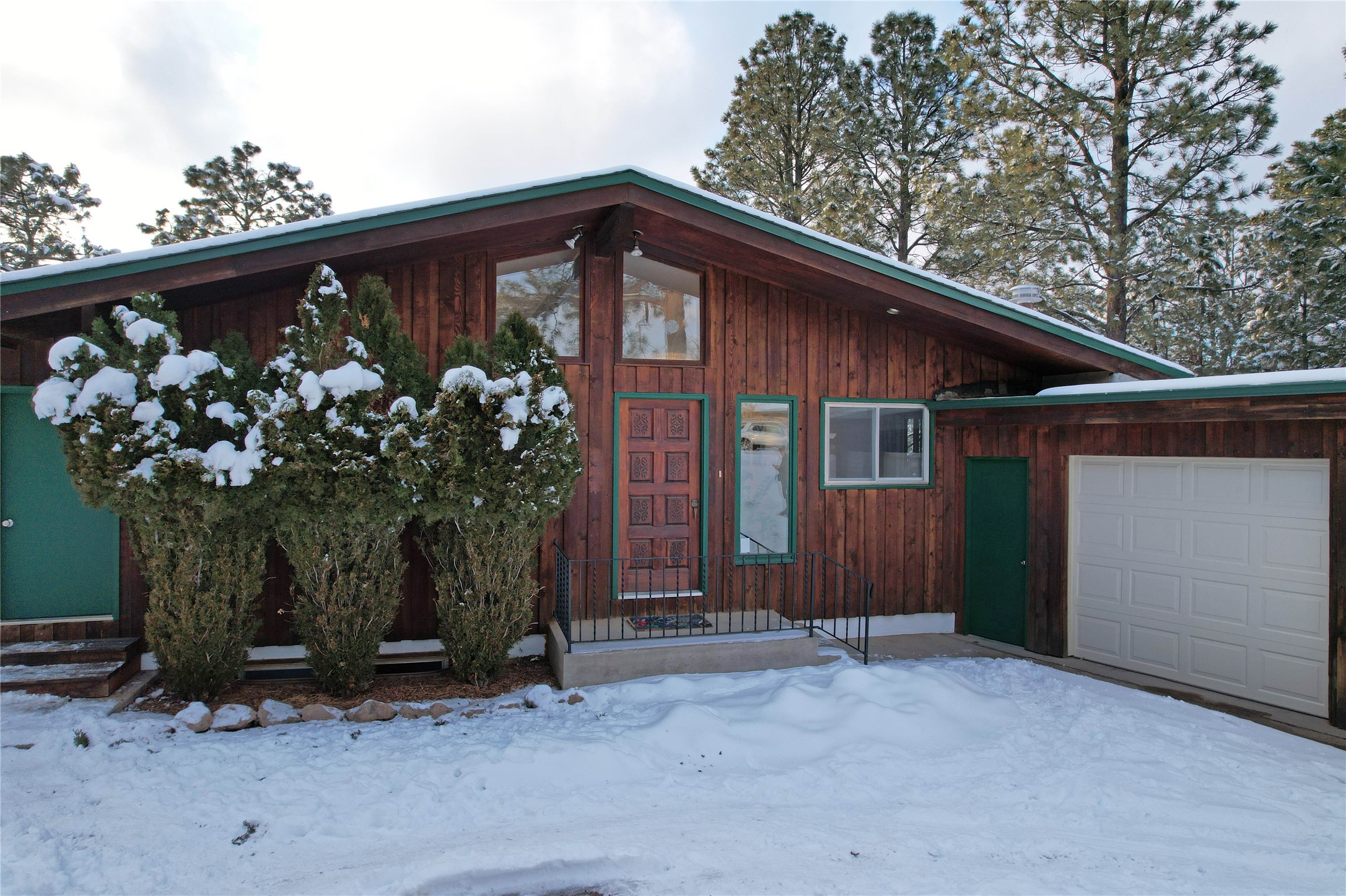 Wood-sided home with green trim and an attached garage, surrounded by snow-covered shrubs and ground.