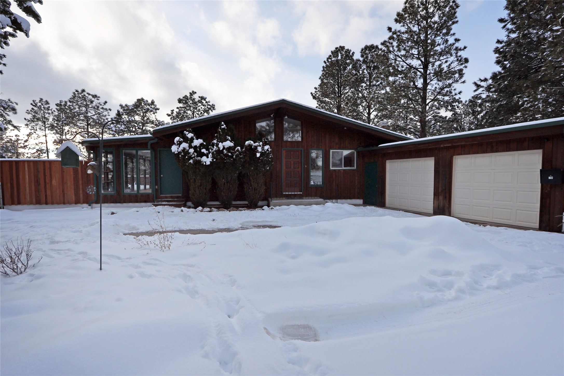 Wood-sided home with two garage doors and a snow-covered yard beneath pine trees.