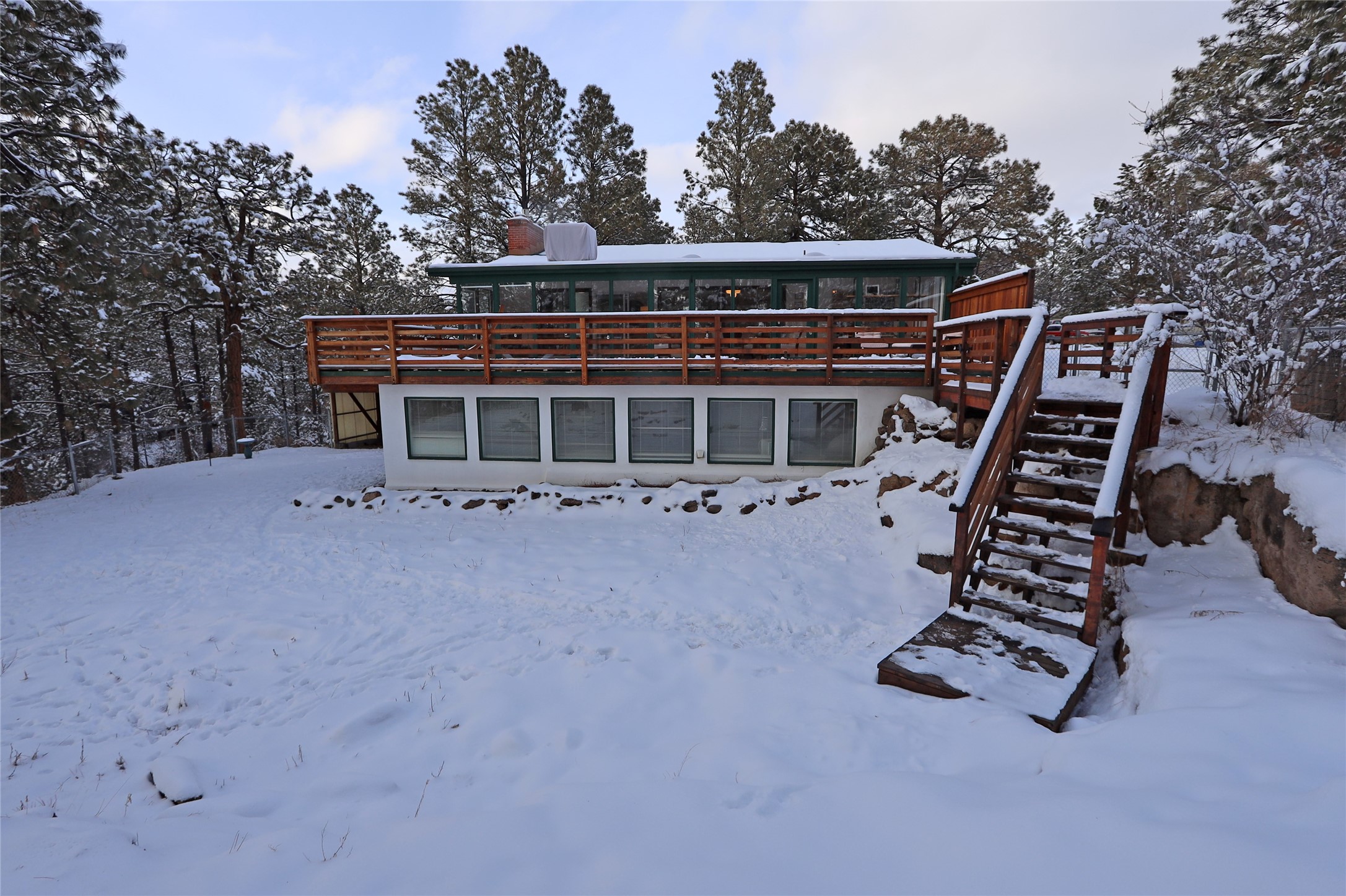 Two-level home with a broad wooden deck and stairs, surrounded by snow-covered ground and pine trees.