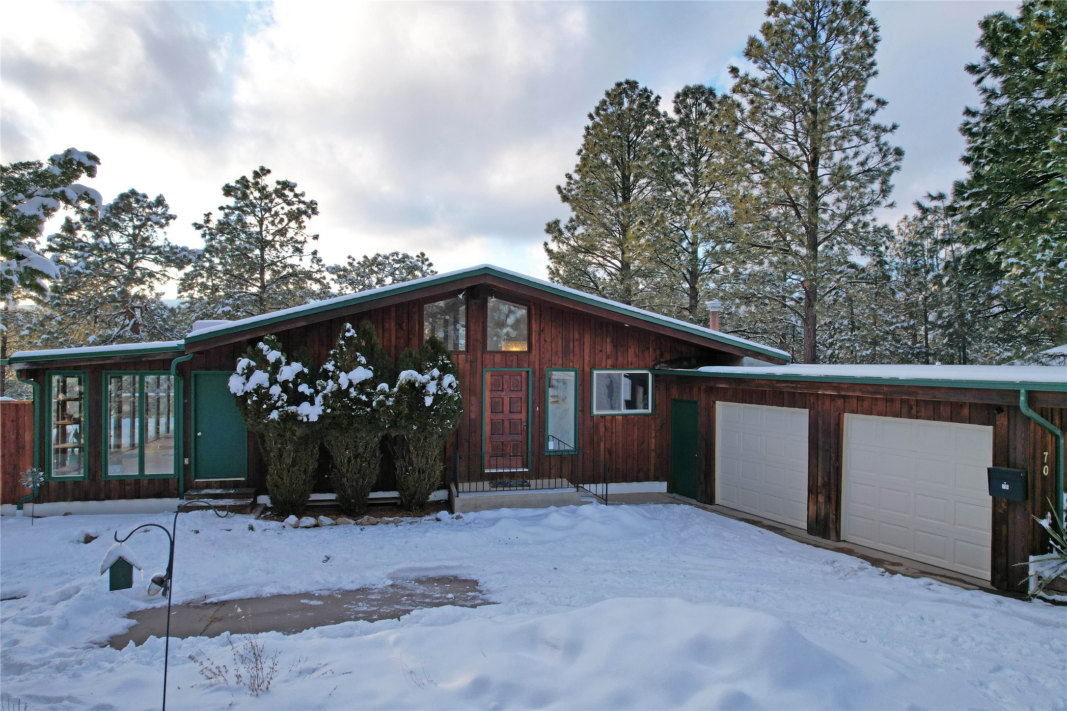 Wood-sided home with green trim and two garage doors, framed by snowy pine trees.