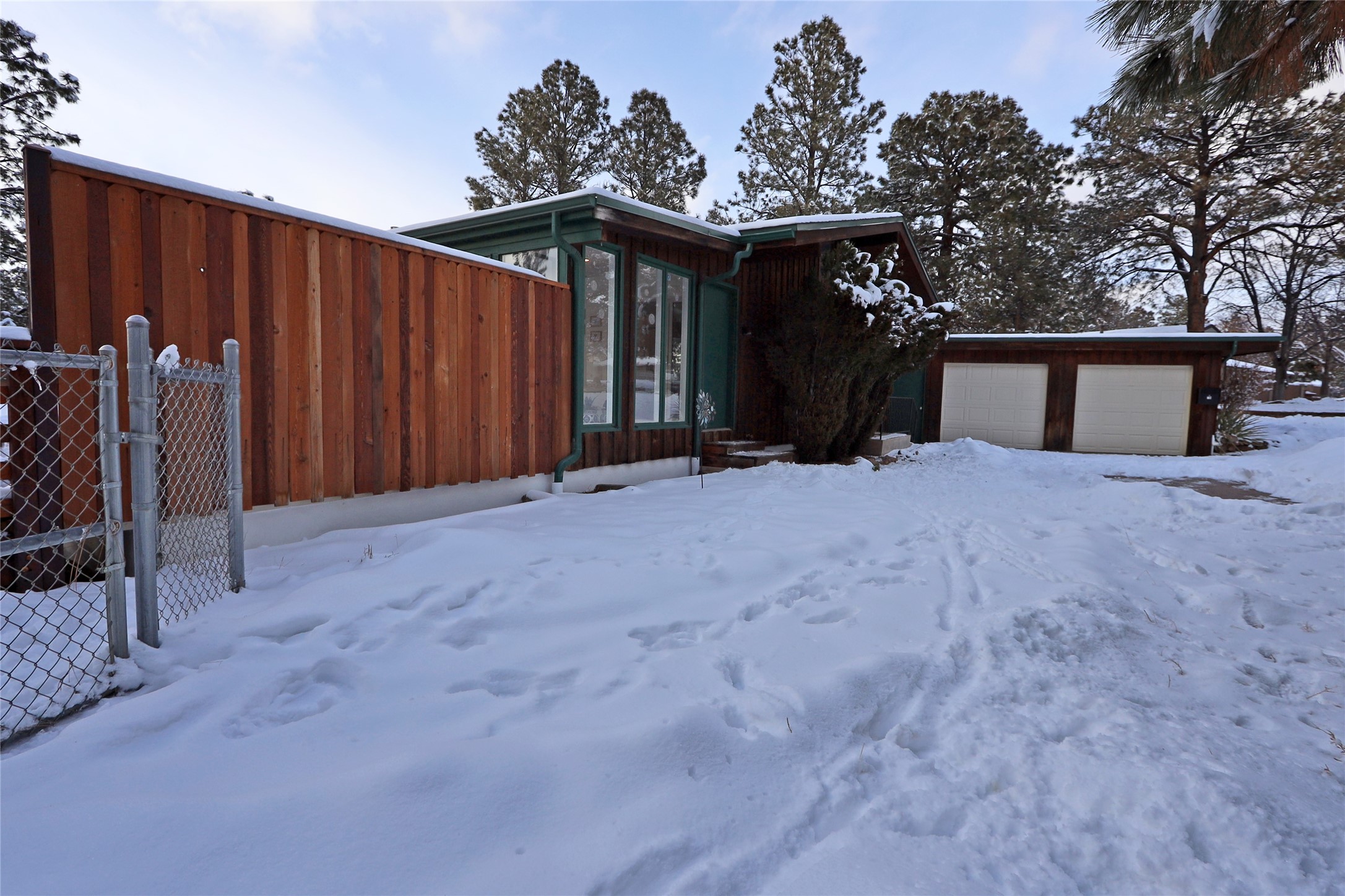 Wood-sided home with green trim and a two-car garage amid snow and evergreen trees.