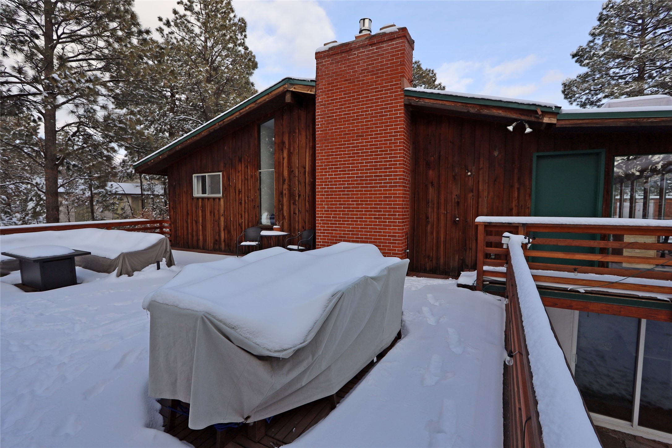 Snow-covered deck with covered patio furniture beside a wood-sided home and tall brick chimney.