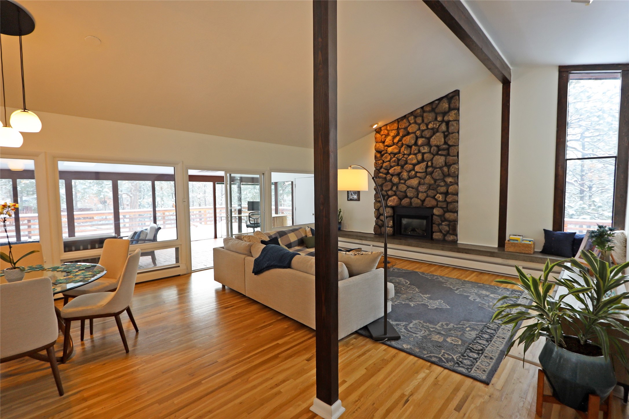 Open-plan living and dining room with a vaulted ceiling, stone fireplace, hardwood floors, and windows overlooking a snowy deck.