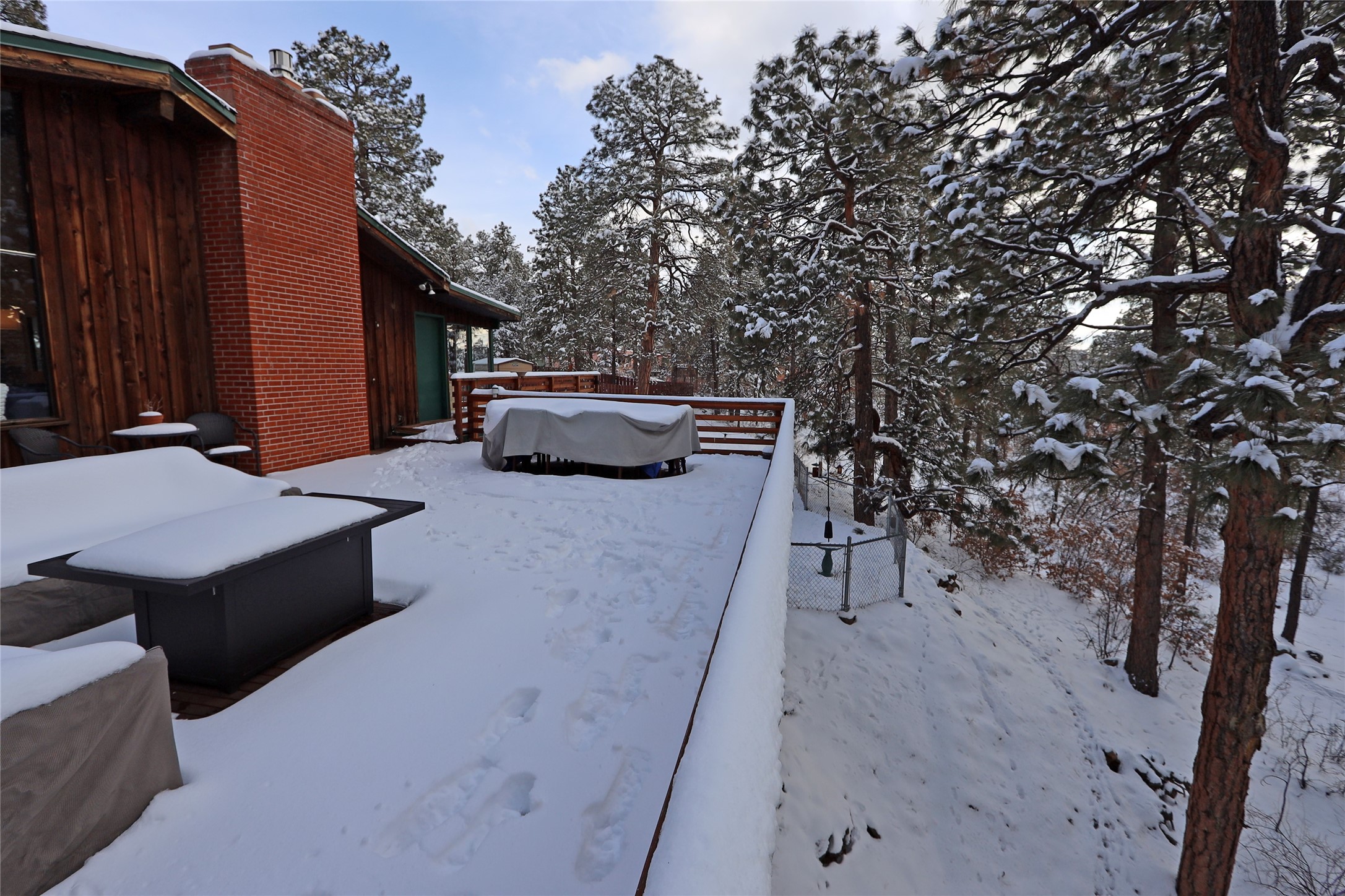 Snow-covered deck with covered patio furniture beside a brick chimney, surrounded by snowy pine trees.