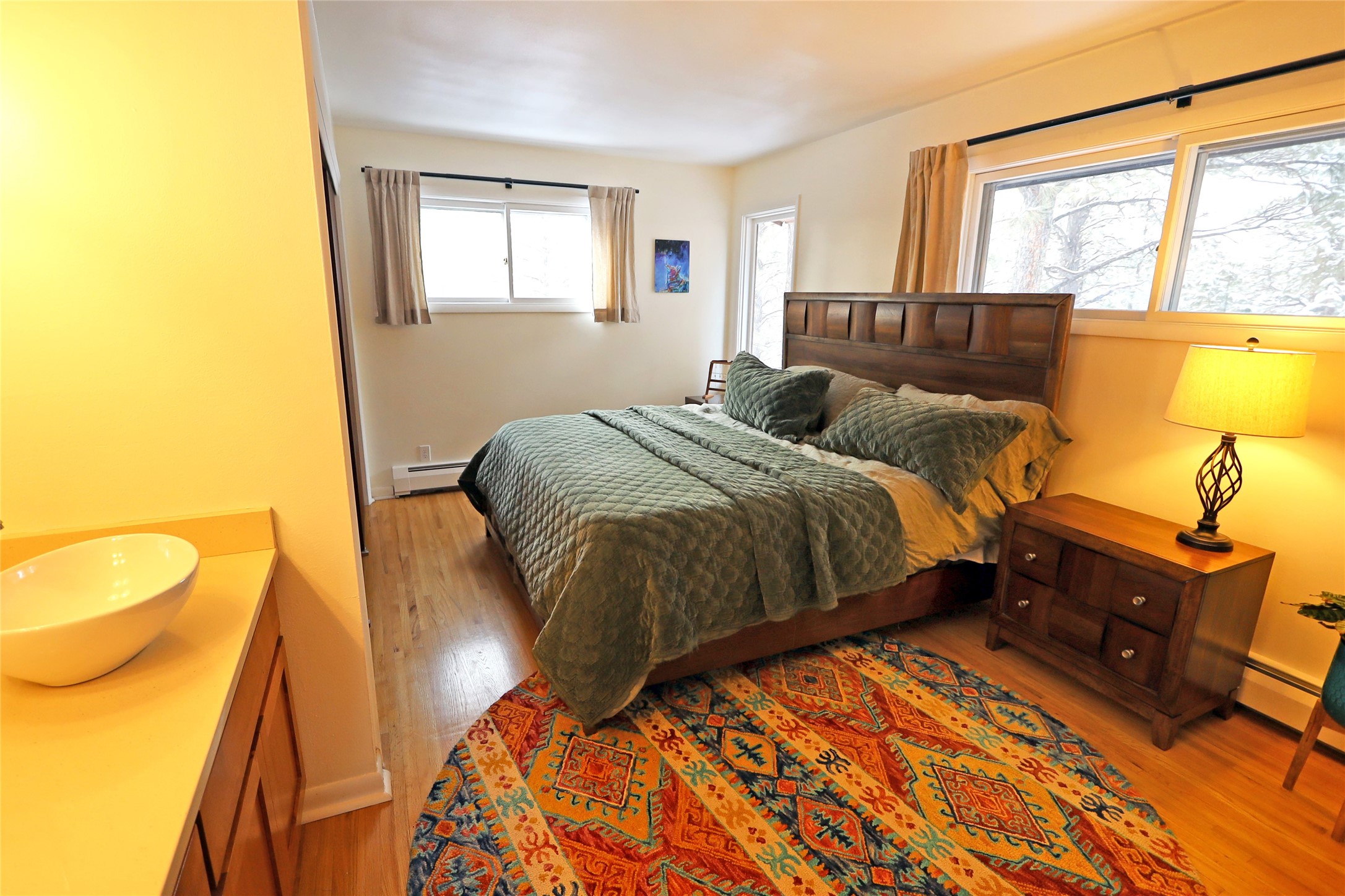 Bedroom with a green-quilted bed, colorful rug, and windows overlooking trees; sink counter in foreground.
