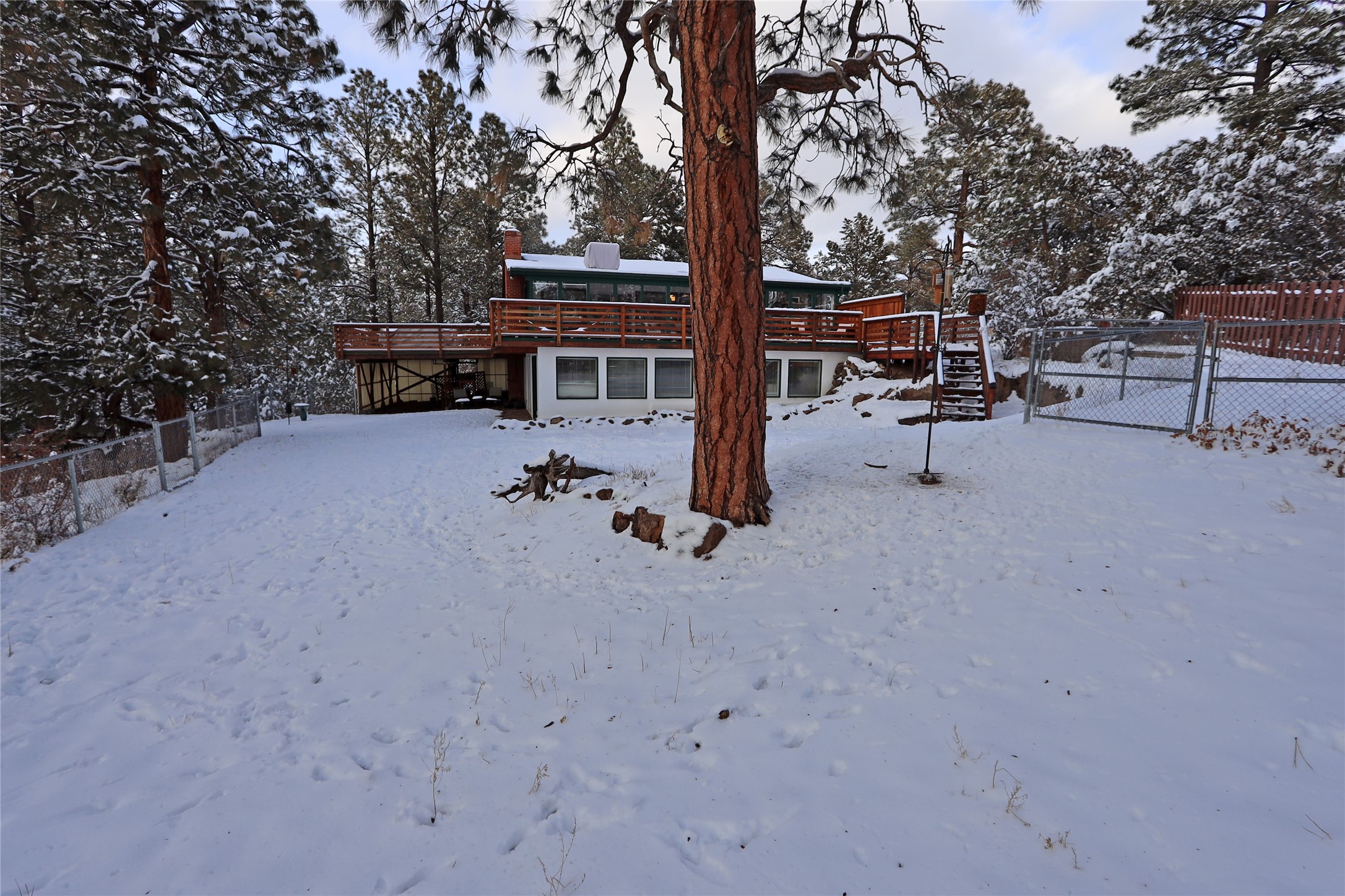 Home with a raised wooden deck and fenced yard amid snow-covered pine trees.