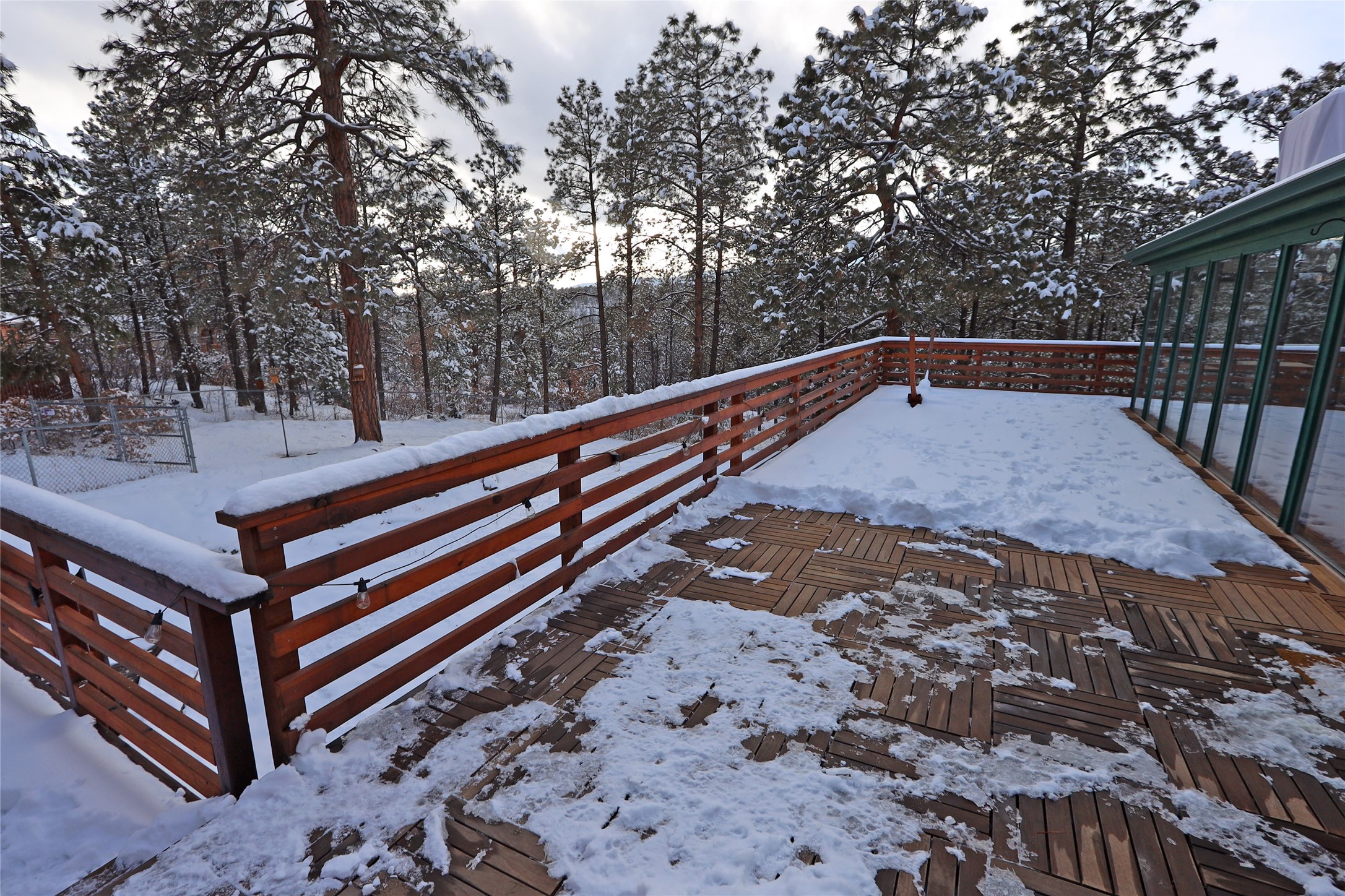 Snow-covered wooden deck with reddish-brown railings, string lights, and pine trees beyond.