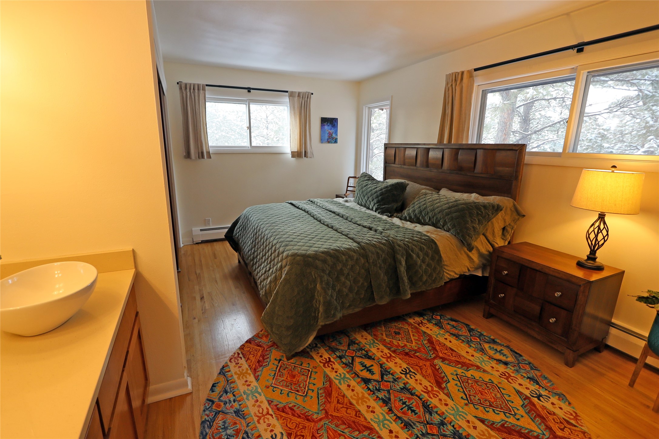 Bedroom with green quilted bedding, a colorful rug, and windows overlooking snow-covered trees.