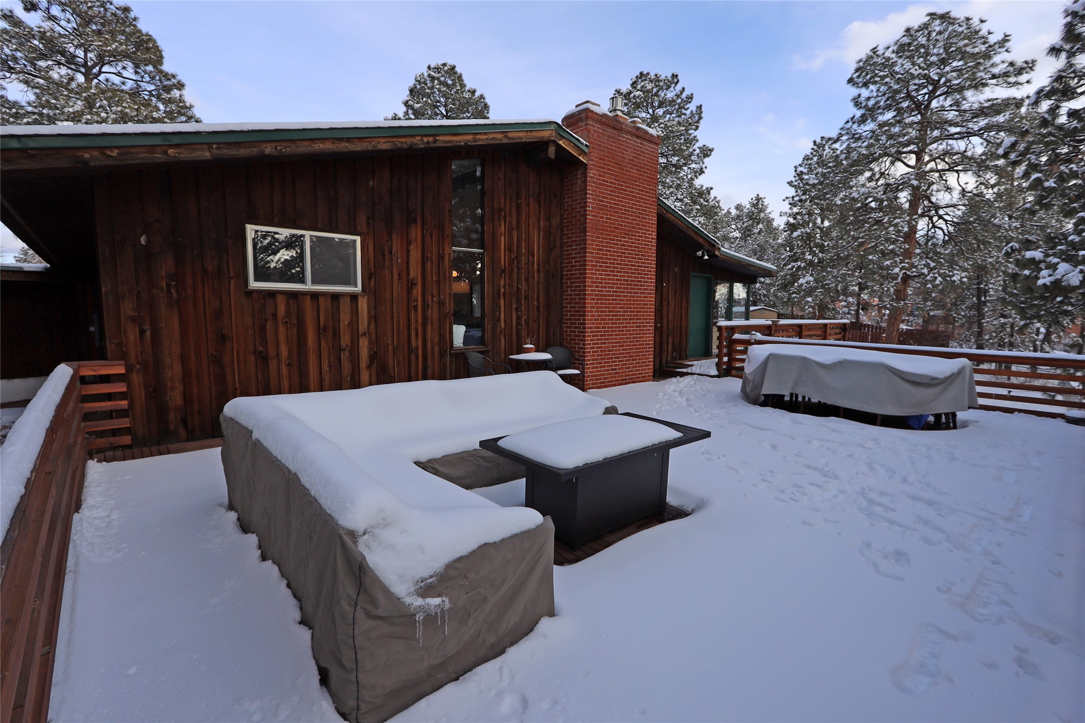 Snow-covered deck beside a wood-sided home with a brick chimney and pine trees.