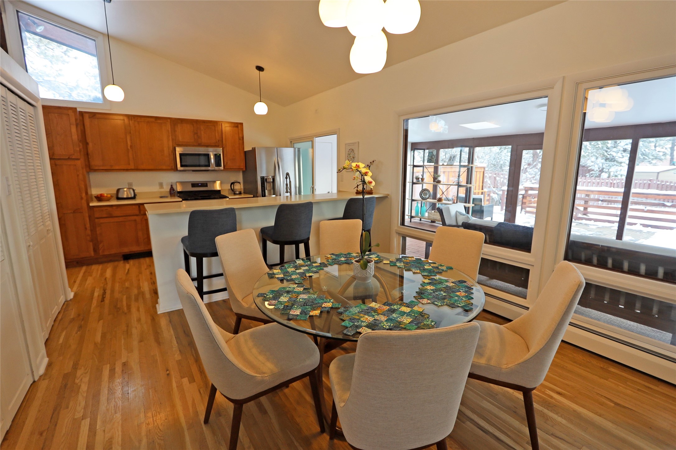 Dining area with glass table and upholstered chairs beside a wood-cabinet kitchen and windows overlooking a snowy deck.