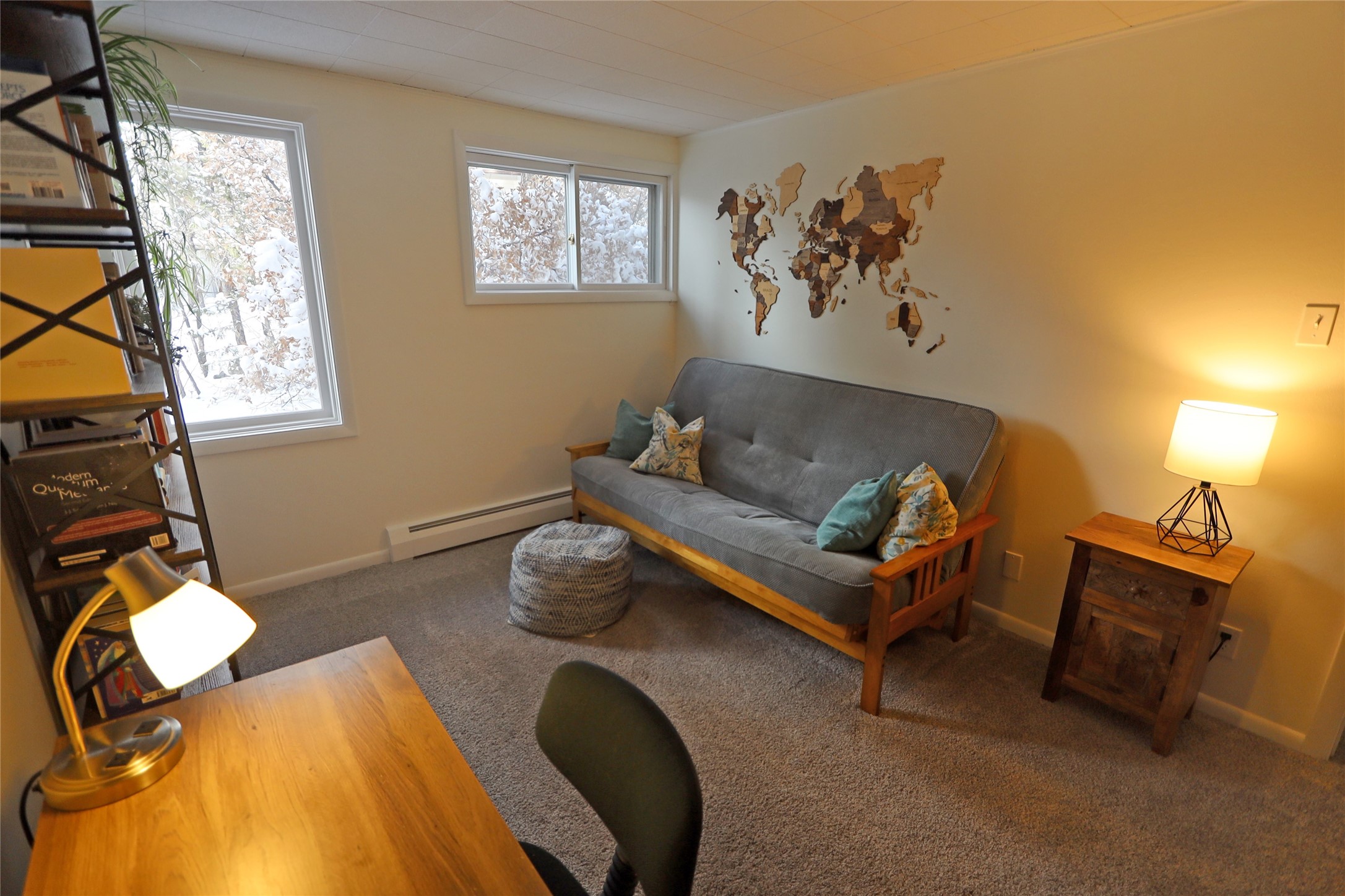 Furnished home office with a gray sofa, wooden desk, wall-mounted world map, and snowy windows.